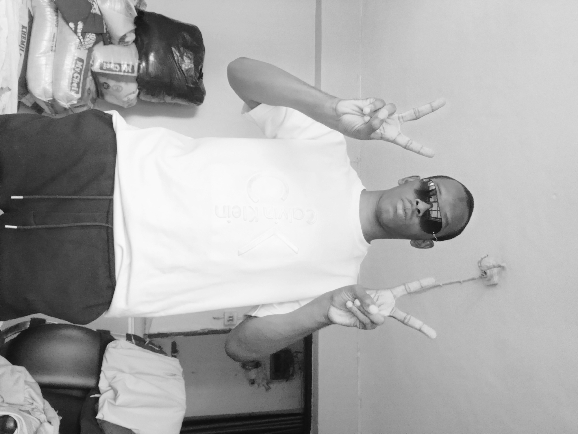 In a medium shot, a Black man stands indoors, posing for a photograph. He wears a white t-shirt with the Calvin Klein logo and black sweatpants. He has sunglasses on and is holding up both hands in a peace sign. The background is dimly lit and cluttered. To his left, there is a stack of bags, some appearing to be filled with rice. To his right, a chair is visible, draped with clothes. The room appears to be a residential space in Lagos, Nigeria.