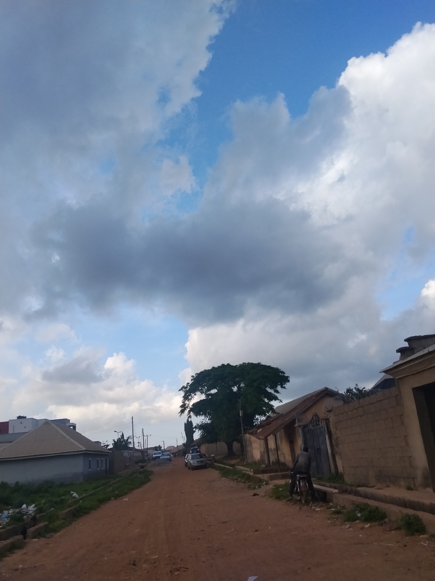 In Mangu Town, Nigeria, a dusty, unpaved road winds through a neighborhood under a partly cloudy sky. The road is lined with buildings and fences made of various materials, including brick and corrugated metal. Greenery, consisting of trees and grass, is visible in patches. On the right side of the road, a person is seated on a motorcycle, facing away from the camera. Further down the road, several cars are parked or in motion. On the left side, a building with a light-colored roof and grey walls stands beside a grassy area with some visible refuse. The overall scene depicts a typical residential street in the town.