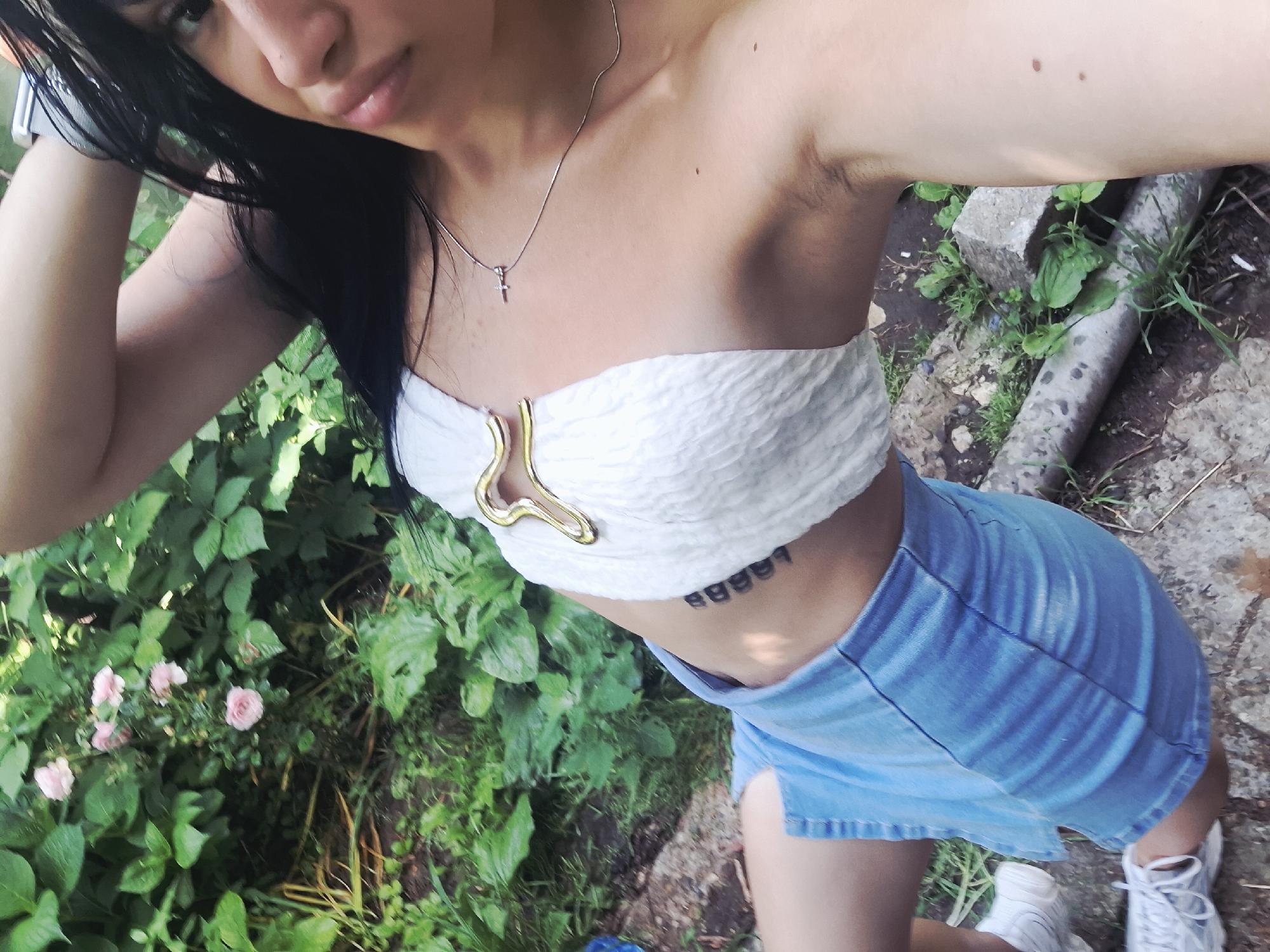 The image is a high-angle selfie of a young woman taken outdoors in what appears to be a garden or overgrown green space. The woman has long, dark hair, fair skin, and is looking slightly upwards towards the camera. She is wearing a white strapless crop top with a distinctive gold-colored, wavy buckle in the center, and a light blue denim mini skirt. A thin silver chain necklace with a small cross pendant adorns her neck. On her right ribcage, a tattoo displaying the characters "88887" is visible. She wears white athletic shoes.
The background is rich with diverse green foliage, including broad leaves and delicate pink roses in bloom towards the lower left. The ground is a mix of dirt, grass, and irregular concrete or stone elements, including what appears to be a concrete pipe or block on the right. The lighting suggests it is daytime. The woman's right arm is raised, likely holding the device used to capture the selfie. Her facial expression is calm. The location context provided is Krapets, Bulgaria.