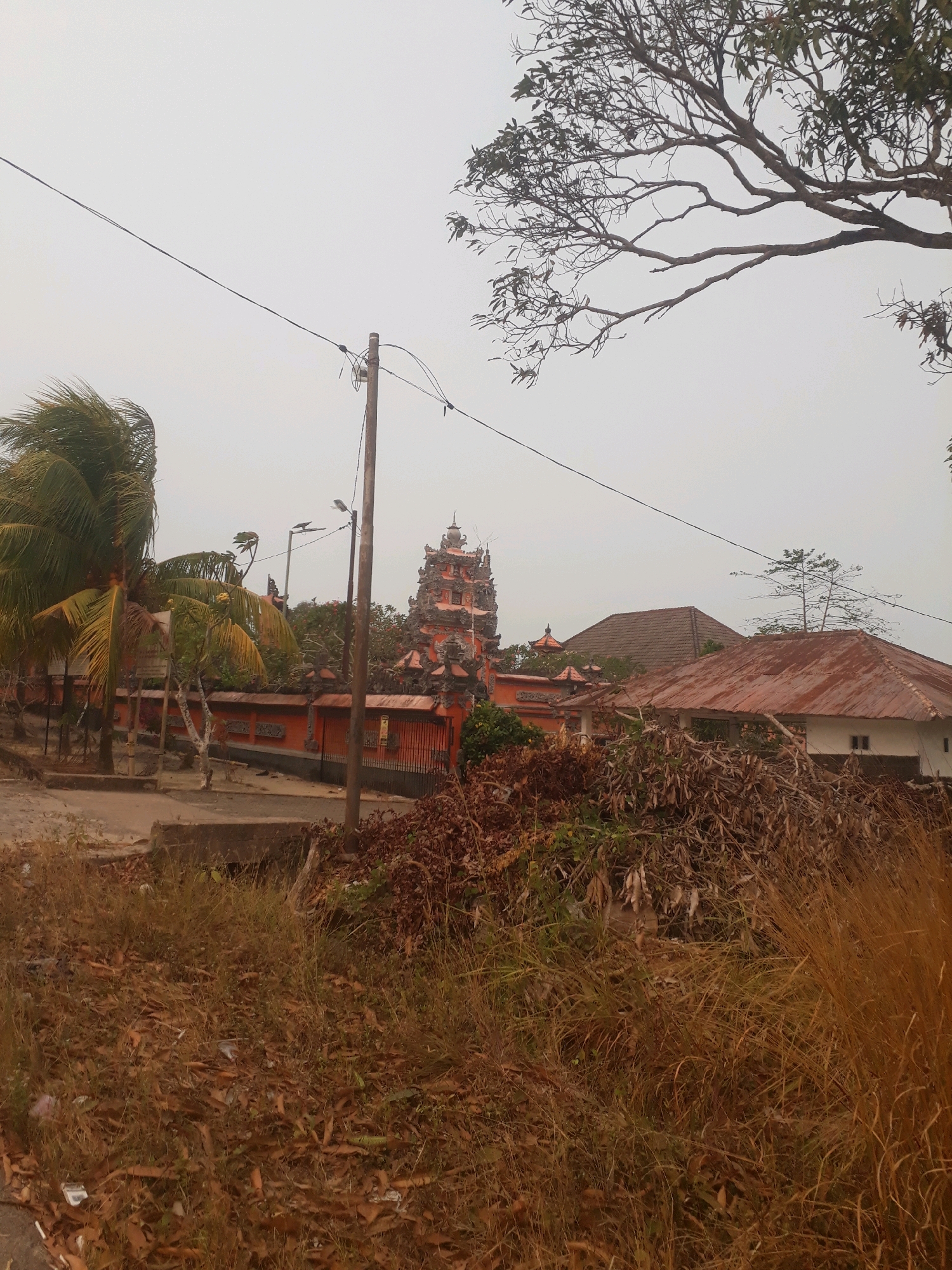 The image depicts an outdoor scene featuring a temple-like structure with orange accents and multiple tiers, resembling Indonesian temple architecture. In the foreground, dry grass and fallen leaves cover the ground, suggesting a natural or perhaps unkempt environment. A utility pole with wires stretches across the frame, and further back, a house with a rusty corrugated metal roof is visible. Palm trees and other foliage frame the scene, indicating a tropical or sub-tropical location. The sky is overcast and hazy, contributing to a subdued atmosphere. The overall impression is of a quiet, possibly rural or semi-urban setting in Pangkalpinang, Indonesia, with a blend of cultural architecture and natural elements.