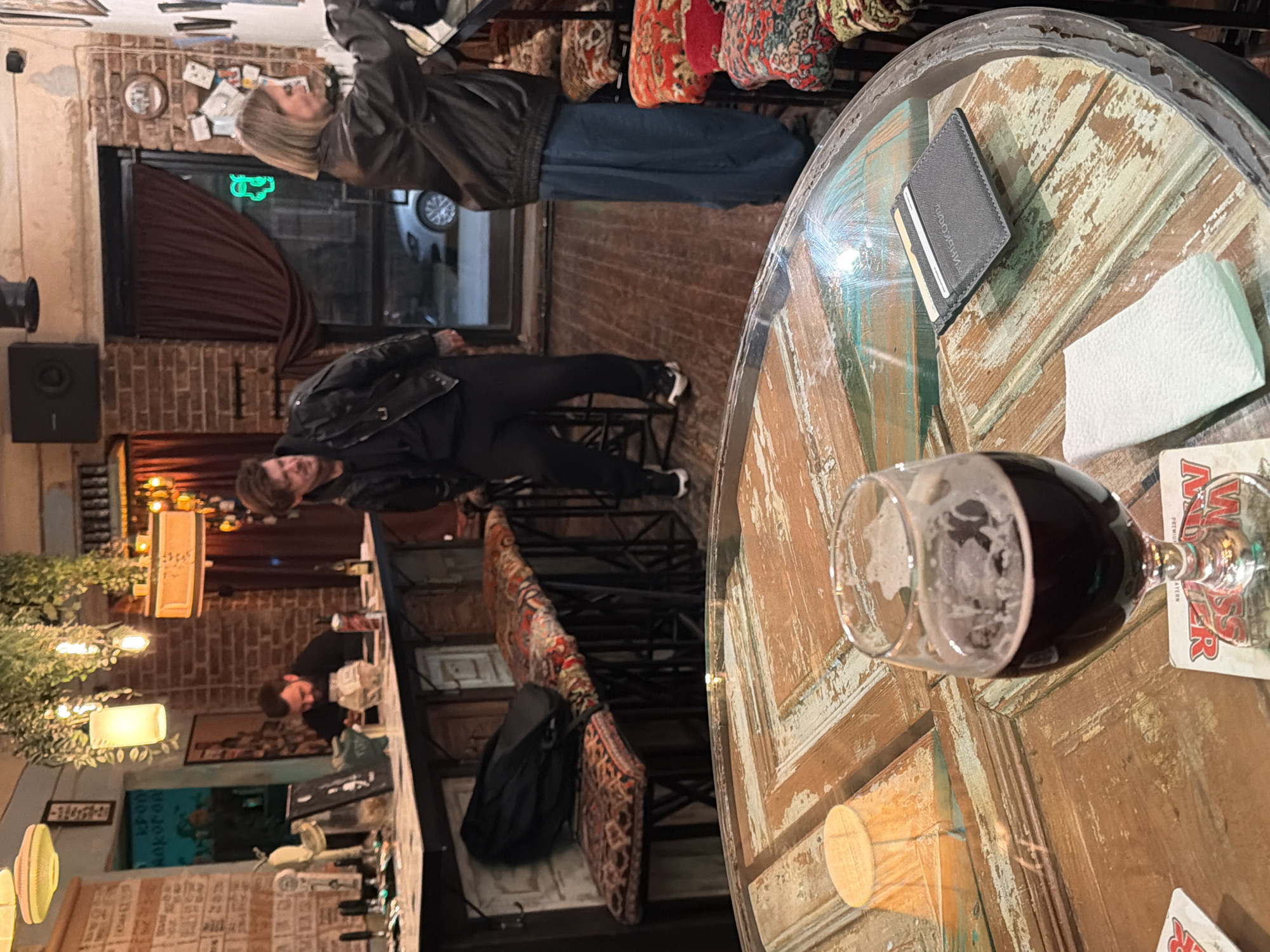 The image shows the interior of a bar, possibly in Rostov-on-Don, Russia, based on the coaster visible on the table.
People: A man and a woman are standing at the bar. Another man is sitting at the bar.
Objects: A round, weathered table with a glass of dark liquid (possibly beer) on it, along with a wallet, a napkin, and a coaster. The bar is lined with stools that have patterned fabric. A backpack is hanging on the back of one of the chairs. There is a beer tap system visible on the bar. There is some vegetation/foliage in the foreground.
Environment: The bar has brick walls. There are decorative objects and lighting fixtures, creating an atmospheric setting. A window is partially obscured by a curtain.