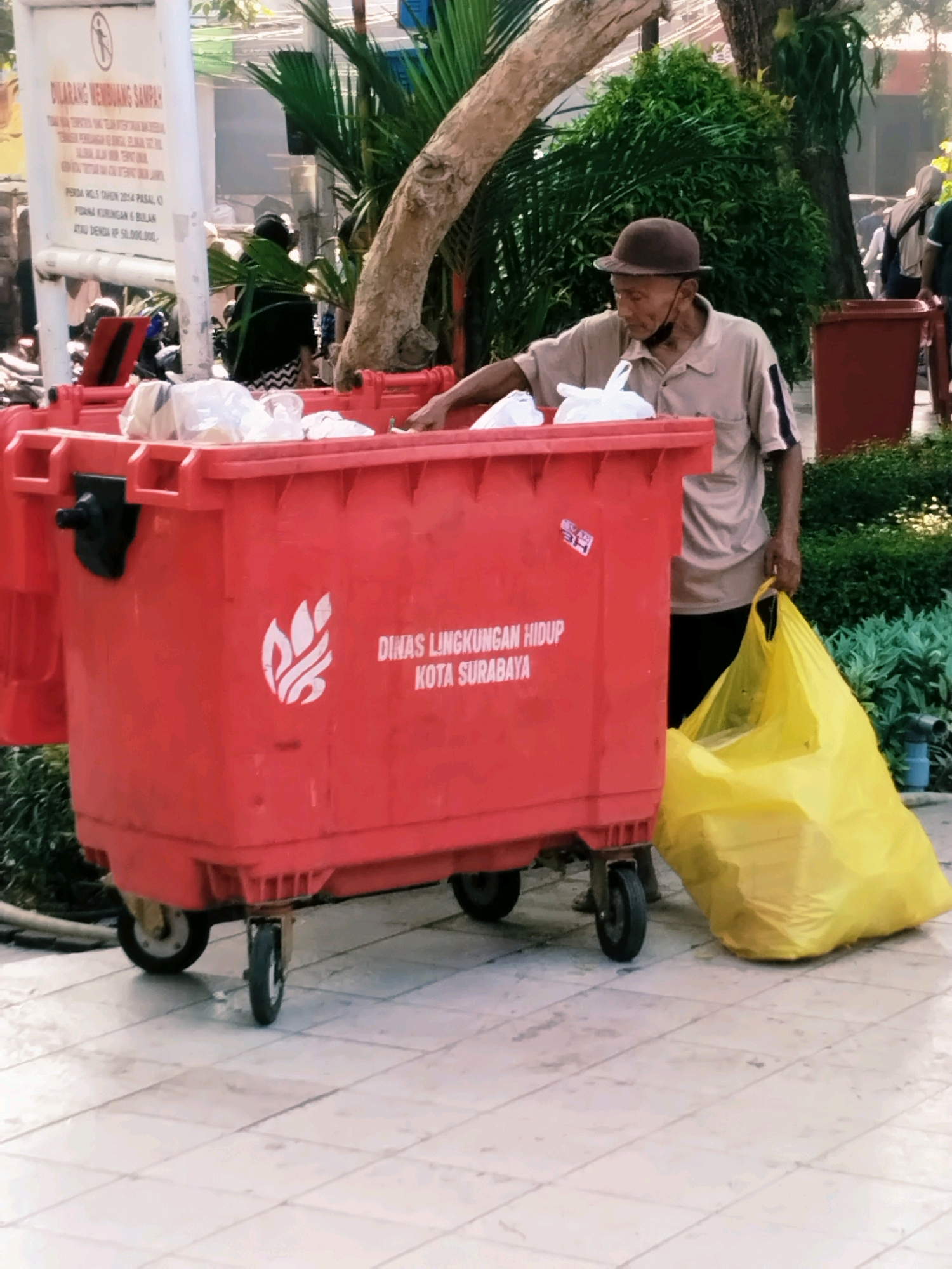 The image shows an outdoor scene with a large red dumpster filled with trash bags. An elderly man wearing a tan polo shirt, a brown hat, and a black face mask tucked under his chin is standing next to the dumpster, holding a large yellow trash bag. He appears to be interacting with the dumpster, possibly depositing the yellow bag. The dumpster has white text and a logo on its side that reads "DINAS LINGKUNGAN HIDUP KOTA SURABAYA," indicating it is part of the Environmental Agency of Surabaya City, Indonesia. The setting appears to be a street or public area with a tiled ground and lush green foliage in the background, suggesting a warm climate. There is also a white sign with text in Indonesian on the left side of the image.