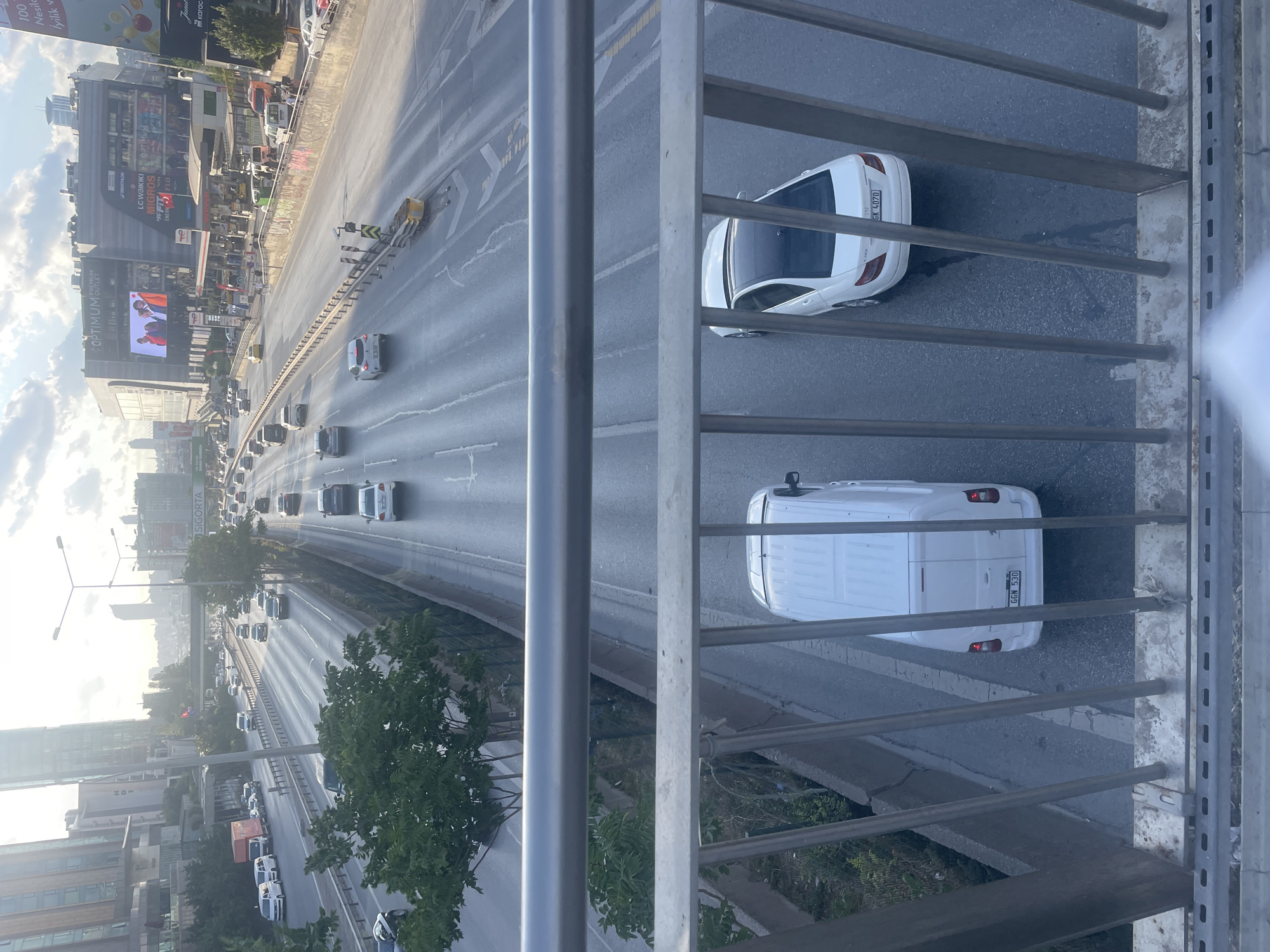 The image, taken from a pedestrian overpass, captures a section of a multi-lane highway in Ataşehir, Turkey, during daylight hours with overcast skies and dappled sunlight. The highway is busy with traffic, featuring a variety of vehicles including cars, vans, and a truck. The infrastructure includes concrete barriers, guardrails, and streetlights. In the background, a modern cityscape is visible, dominated by tall buildings, including a prominent shopping center named "OPTIMUM OUTLET" with a large digital display screen and commercial signage. The overpass itself is constructed of metal, with vertical bars and horizontal railings forming a barrier between the viewer and the scene below. The road surface appears wet, suggesting recent rain.