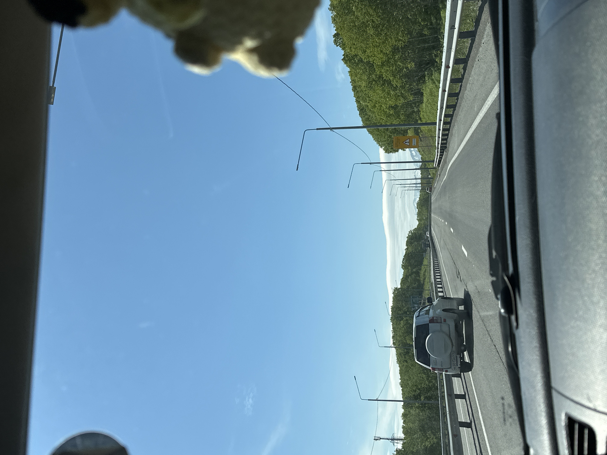 The image is taken from the perspective of a driver inside a car on a highway. The road ahead is clear, with a white SUV in front and a few other vehicles in the distance. The highway is lined with trees and lampposts. The sky is blue with a few scattered clouds. The location is identified as being in the city of Svetly, Russia, specifically on a highway. There are no people visible outside the vehicles. The overall scene depicts a typical day on a highway in a rural or semi-rural area.