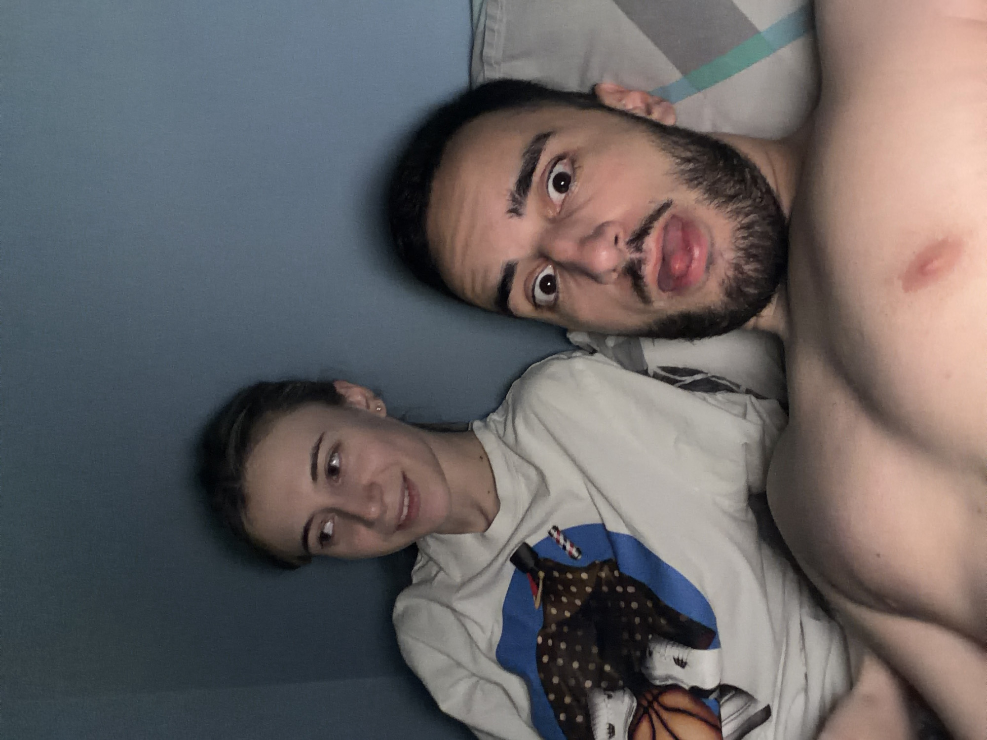 A man and a woman are lying in bed. The man, shirtless with a beard, has a surprised expression. The woman is wearing a t-shirt with a graphic print. They are likely indoors. The image was not taken in the city of Plovdiv, Bulgaria.