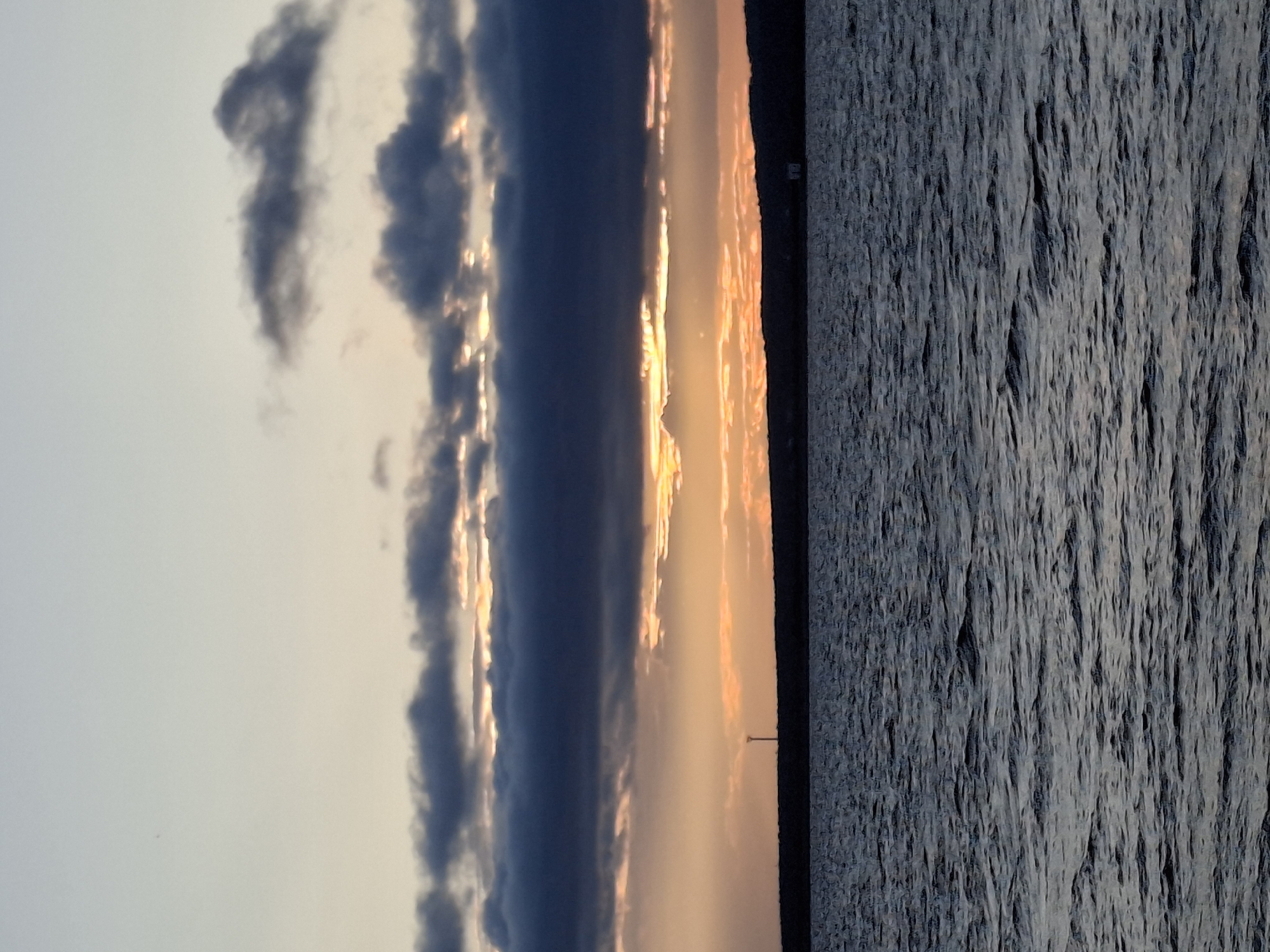 The image depicts a serene body of water, likely a reservoir or river, under a cloudy sky during either sunrise or sunset. The water's surface is rippled, reflecting the muted colors of the sky. A dark, low-lying horizon line, possibly a shoreline with trees or hills, separates the water from the sky. A thin, tall structure resembling a lamppost or marker is visible on the horizon on the left side of the image. In the distance on the right, a small, light-colored building or structure can be observed. The sky is characterized by layers of clouds, with some sections illuminated by warm, orange and yellow light, suggesting the sun is either rising or setting. The overall impression is one of natural beauty and tranquility.