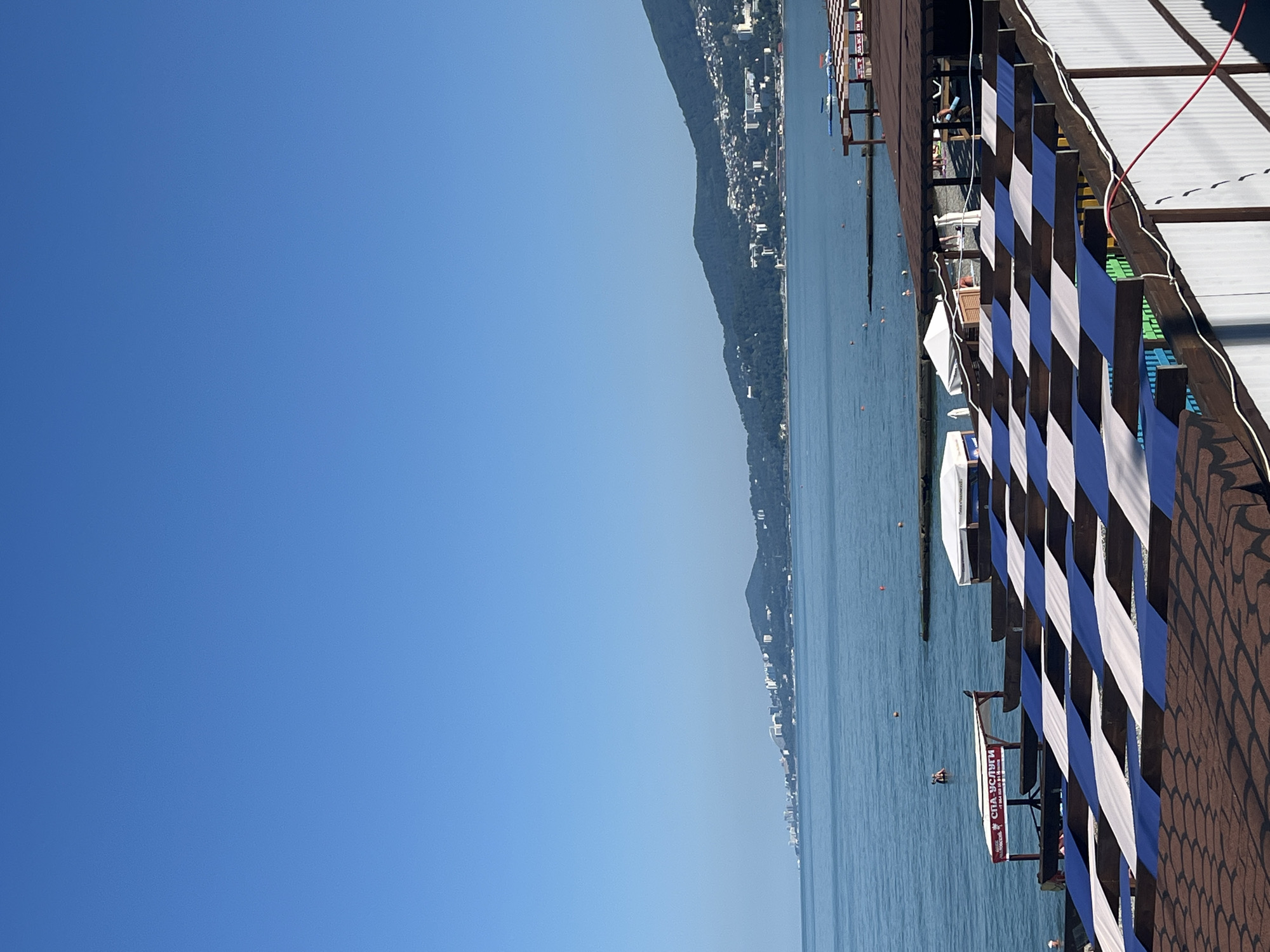 This image captures a sun-drenched beach scene in Sochi, Russia, under a clear blue sky. In the foreground, a row of beach umbrellas with alternating blue and white panels shades a seating area. Beyond this, the calm, blue sea stretches to the horizon, meeting a mountainous coastline dotted with buildings. Several people are visible in the water, some swimming, while others relax on the beach or under shaded structures. A red sign reading "СПА-УСЛУГИ" (SPA SERVICES) is prominently displayed near the water's edge. The overall atmosphere is one of a typical summer day at a seaside resort.