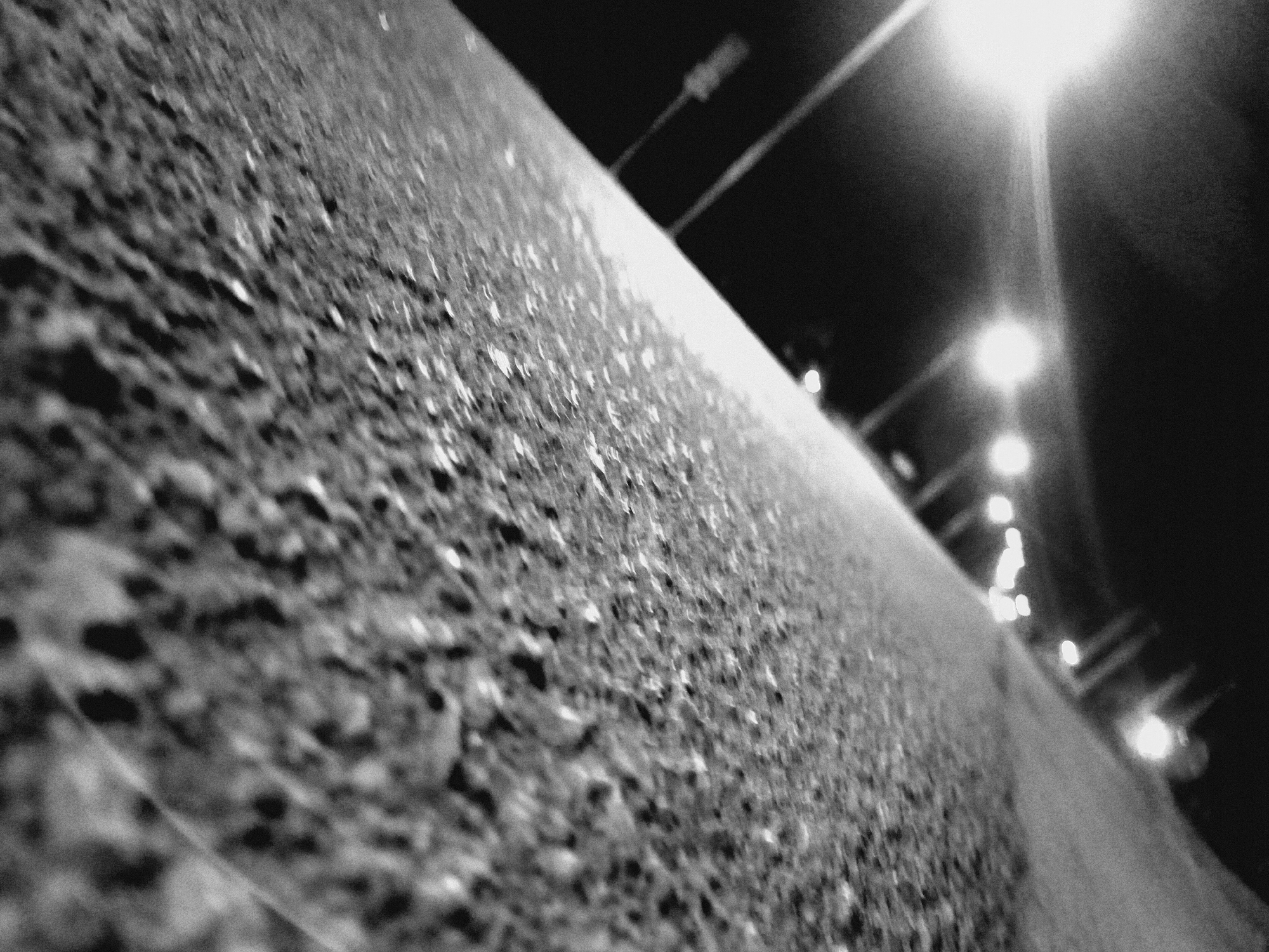 A black and white photograph taken at night, possibly in Barakin Angundi, Nigeria, shows an extreme close-up of rough asphalt or concrete. The perspective is low and angled, making the surface appear to extend into the distance. Streetlights are visible in the upper right portion of the image, blurred and out of focus due to the depth of field. Their poles are silhouetted against the dark sky. The focus is concentrated on the texture of the surface, showing its irregular pattern of bumps and pits.