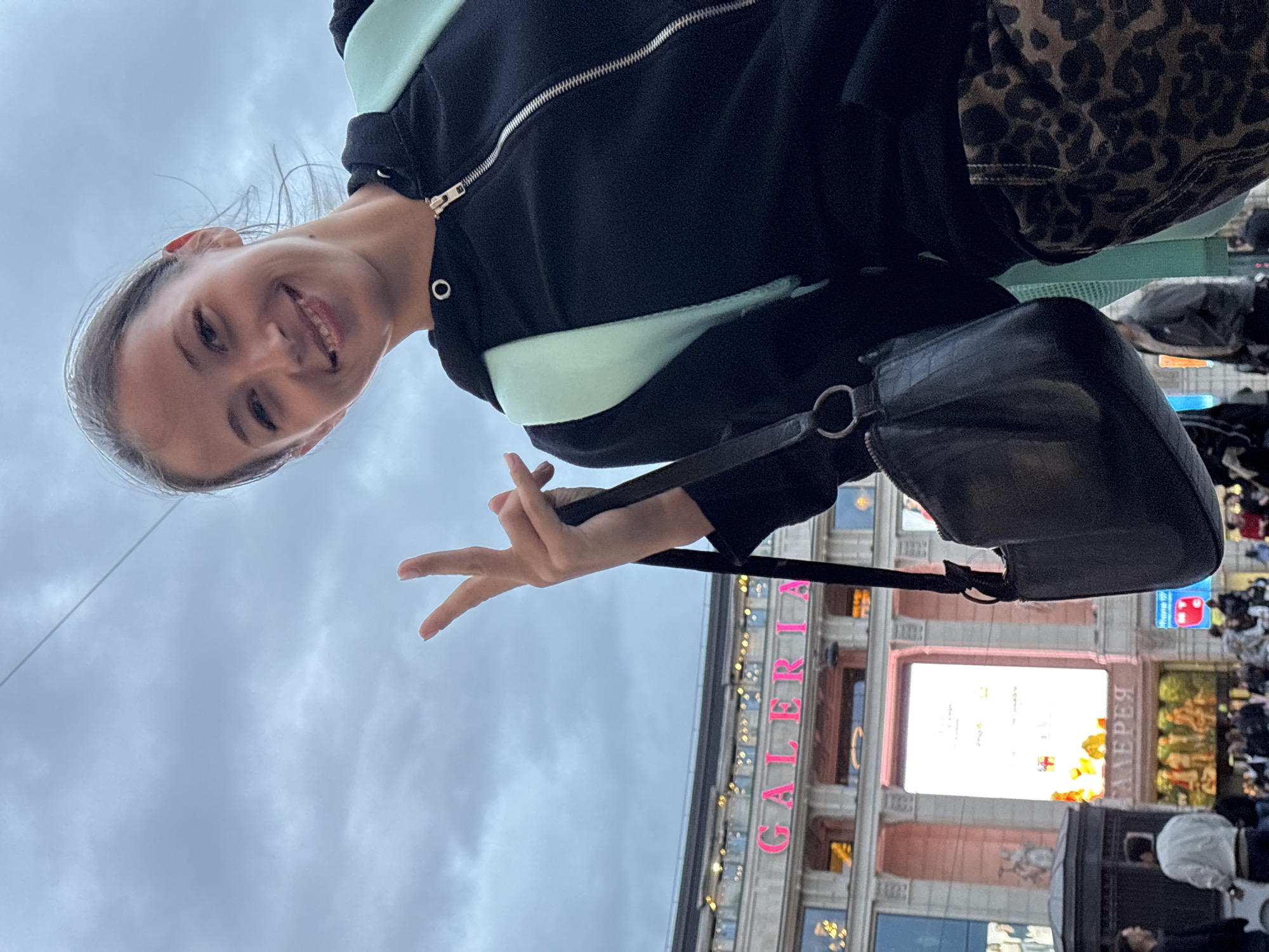 A woman smiles at the camera, holding up two fingers in a peace sign. She wears a black and light green hooded sweatshirt. A black purse hangs on her shoulder. In the background is the "Галерея" mall in Saint Petersburg, Russia. There are people in the distance. The sky is overcast.