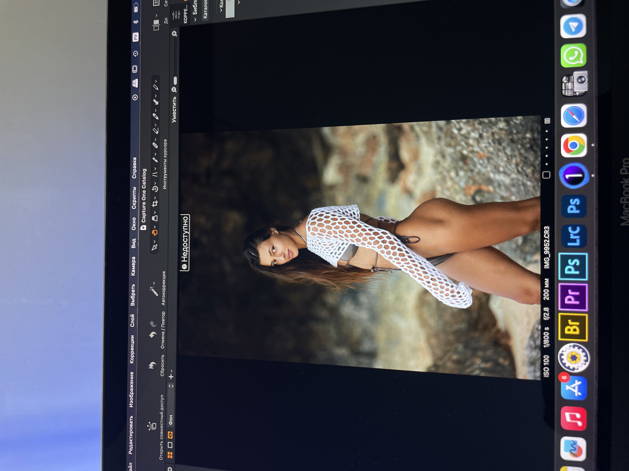 The image shows a photograph of a woman with dark hair wearing a white crocheted top and a bikini, posing against a rocky backdrop. It appears to be a shot from a photo editing program on a MacBook Pro. The program, Capture One Catalog, is open, with various editing options visible. The bottom right of the screen shows icons for other Adobe programs such as Photoshop, Lightroom and Premiere Pro, along with other Mac apps. The image was taken at ISO 100, 1/800 s, f/2.8, and 200 mm. The location of the shoot is not visible, so it is impossible to know if the photo was taken in Kazan, Russia.