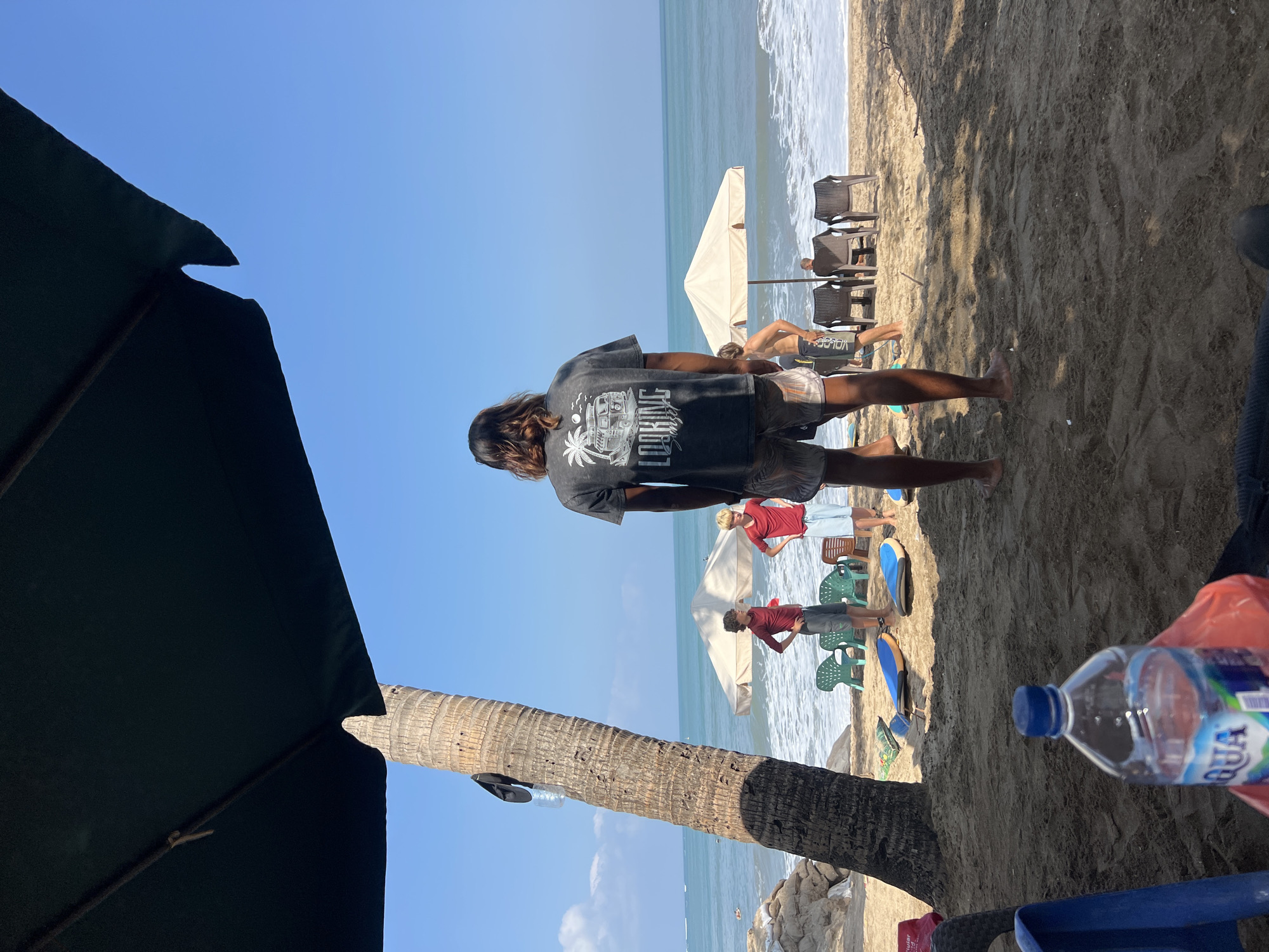 An outdoor, eye-level shot captures a beach scene in Kuta, Indonesia on a sunny day. In the foreground, a person with long hair stands with their back to the camera, wearing a dark t-shirt with a graphic print and patterned shorts. Their feet are bare. To the left, a large, dark umbrella shades a portion of the sandy ground. A coconut tree trunk is visible to the left of the umbrella.
In the middle ground, several people are present. Two young men, one in a red t-shirt and grey shorts, and another in a red t-shirt and white shorts, stand on the sand. A third younger man, in a red long-sleeved shirt and grey shorts, stands nearby. Further back, other individuals are visible near beach chairs and umbrellas. Surfboards are scattered on the sand in front of some of the people.
The background features the ocean with gentle waves breaking on the shore. The sky is clear and blue. The sand on the beach is a light brown color, with shadows cast by the umbrella and palm tree. A few plastic chairs and beach umbrellas are set up on the sand. A plastic bottle of "AQUA" brand water is in the lower foreground.
