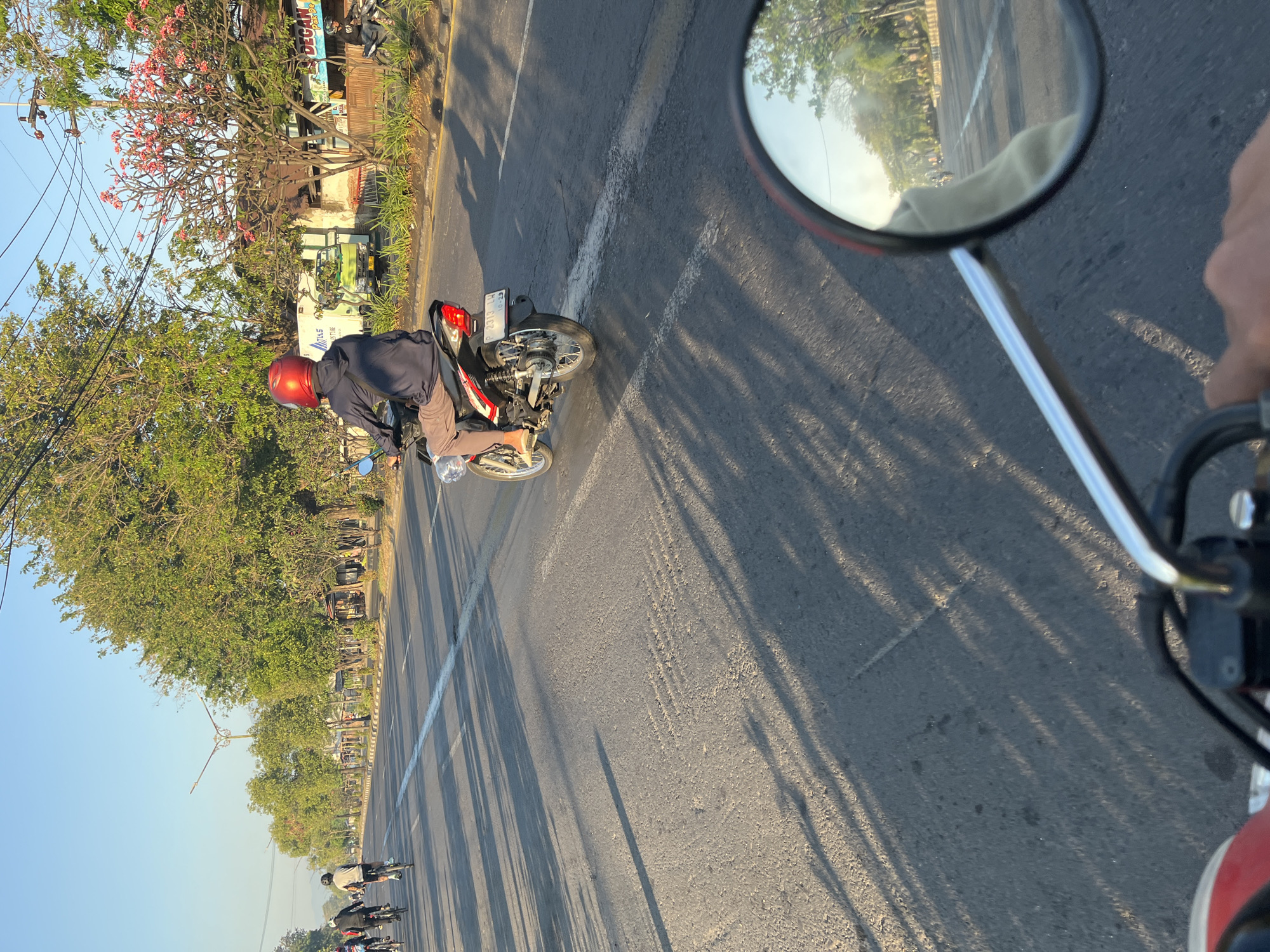 The image depicts a street scene in Ketapang, Indonesia, bathed in the golden light of early morning or late afternoon. A person wearing a red helmet is riding a motorcycle down a paved road, with other cyclists visible in the distance on the left side of the frame. The road is lined with trees and buildings, suggesting an urban or semi-urban environment. A white truck with "INT" visible on its side is parked on the right side of the road. The foreground of the image shows a motorcycle's rearview mirror, reflecting the scene ahead and to the side, including trees and a road. The overall atmosphere is one of daily life and transit.