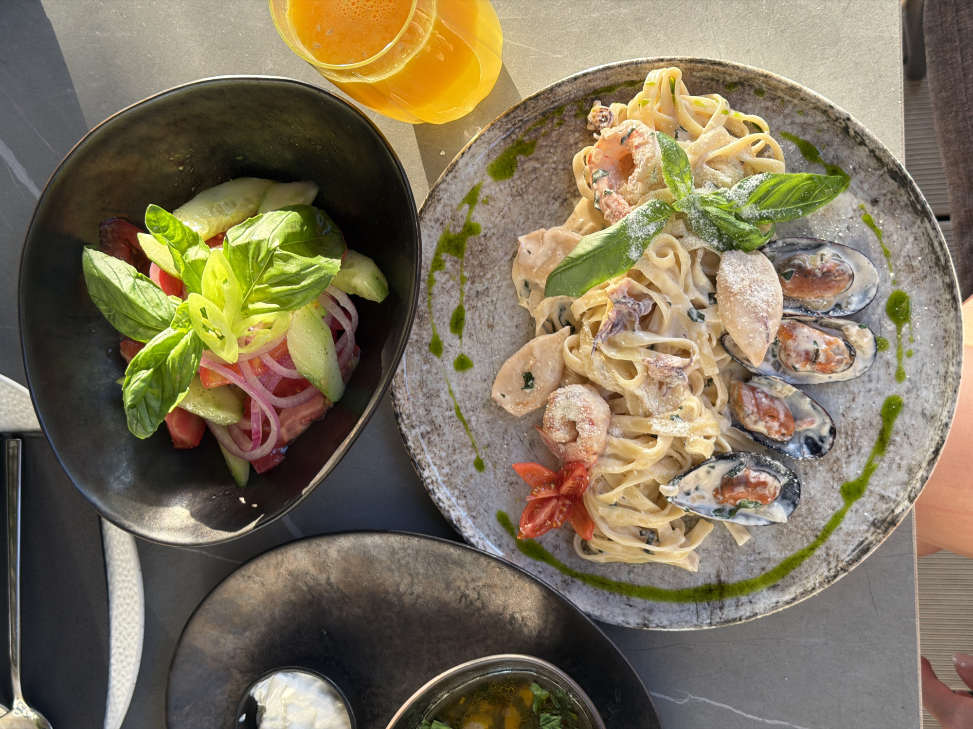 A top-down view shows a meal laid out on a grey surface, likely a table. The scene could plausibly be located in a restaurant in Rafailovići, Montenegro. The meal consists of three main components: a salad, a pasta dish with seafood, and a glass of orange juice.
The salad is in a dark-colored bowl and appears to contain tomatoes, cucumbers, red onions, and basil. The pasta dish, served on a speckled grey plate, features fettuccine-style pasta in a creamy sauce with shrimp, squid, and mussels. A small flower made from a tomato slice and basil leaves garnish it. The glass of orange juice is clear and filled with an orange liquid.
Beneath the pasta plate and to the side of the salad bowl, a darker plate is visible, with a small bowl containing a creamy white substance and another bowl filled with a green liquid. A silver fork is visible to the left.
A small portion of a person's upper arm and hand is visible in the right side of the image.