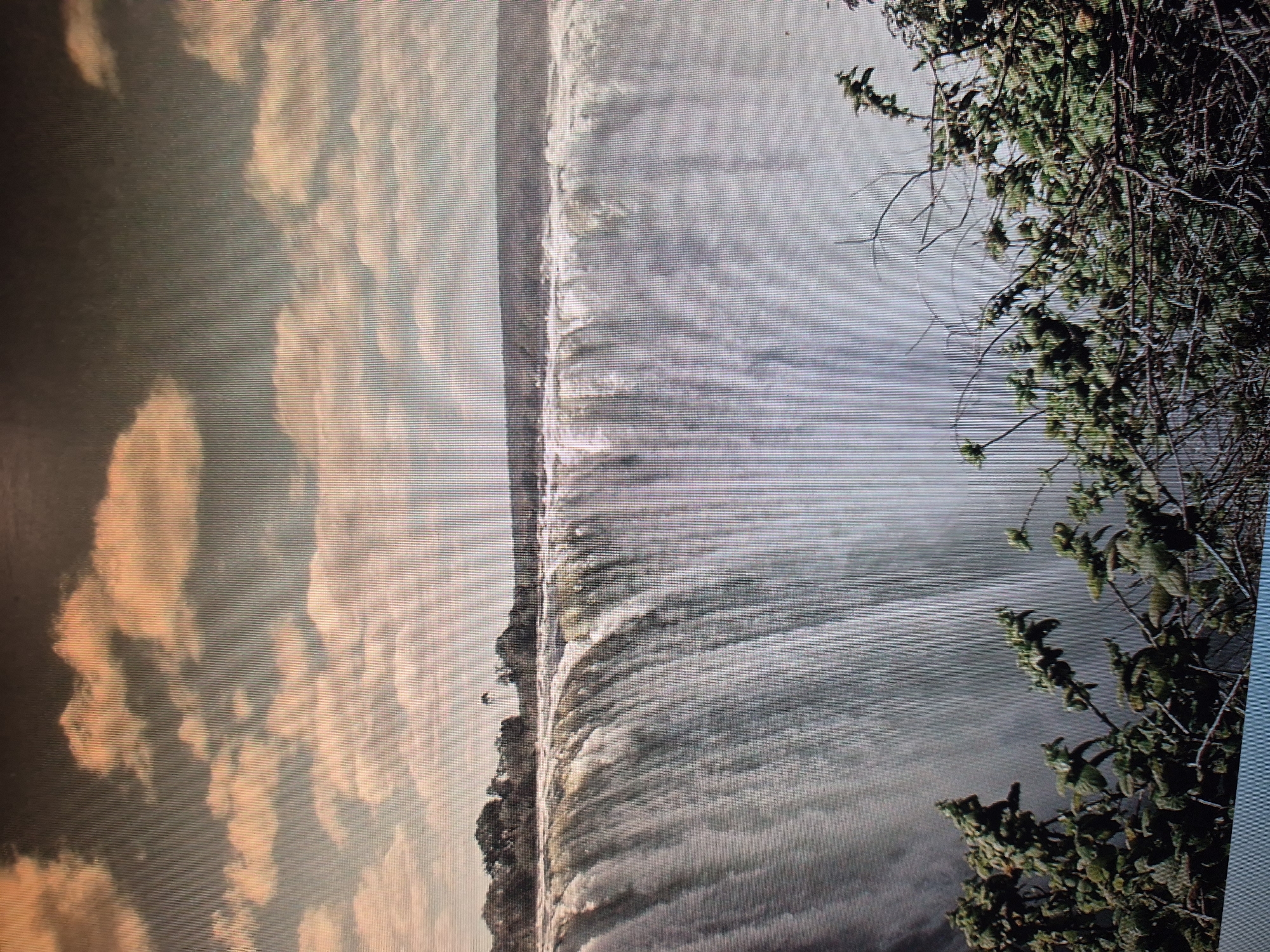 The image shows a large waterfall with water cascading over a cliff edge. On the left side of the frame, there are clouds in the sky with a slightly reddish hue, possibly indicating sunrise or sunset. Vegetation, including trees and bushes, is visible along the top of the cliff and on the right side of the frame. The water appears turbulent and creates mist as it falls. There is no evidence to suggest this photo was taken in the city of Yerevan, Armenia.