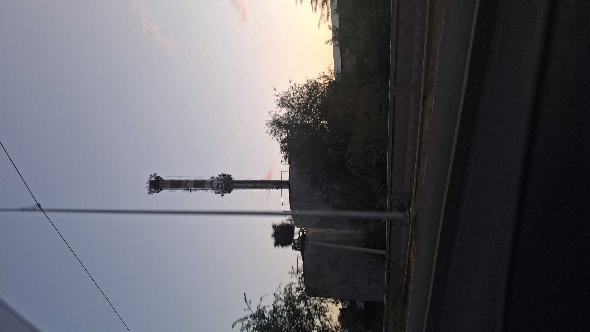 The image shows a communication tower situated between two large, cylindrical concrete structures in an industrial or semi-industrial area, possibly near Shymkent, Kazakhstan. The tower is tall and slender, with multiple antennas and communication equipment visible at its apex. The concrete structures appear to be water reservoirs or storage tanks, with one having a dark, irregular object attached to its side. Dense foliage surrounds the base of the tower and the concrete structures, suggesting a natural or semi-natural encroachment on the industrial site. A grey concrete barrier runs across the foreground, separating the area from a dark asphalt road. The sky above is a gradient of pale blue to soft orange, indicating either sunrise or sunset. A thin black cable stretches diagonally across the upper portion of the frame. The overall scene depicts a juxtaposition of technological infrastructure and natural elements under a twilight sky.
