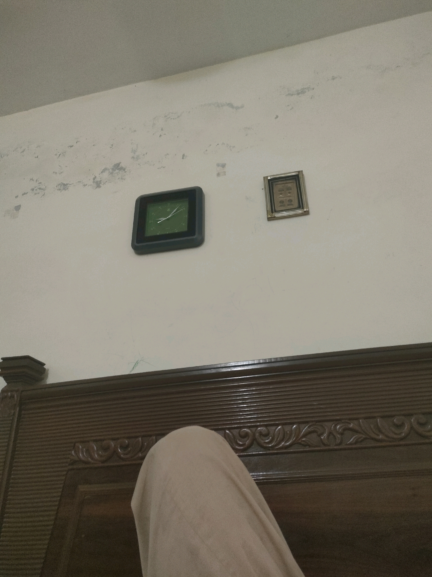 The image shows a close-up view of a bedroom interior. On a textured, off-white wall, there are two objects. A square, dark-framed clock with a green face and white hands is positioned on the left. To its right, a smaller, square decorative item with a metallic frame, containing four circular motifs, is also mounted on the wall. Below these objects, the top portion of a wooden headboard is visible. The headboard has horizontal grooved detailing and ornate carved elements. A section of light-colored fabric, possibly bedding or clothing, is draped over the right side of the headboard, obscuring part of the view. The lighting suggests an indoor environment, likely during the day. The scene is located in Tandlianwala, Pakistan.