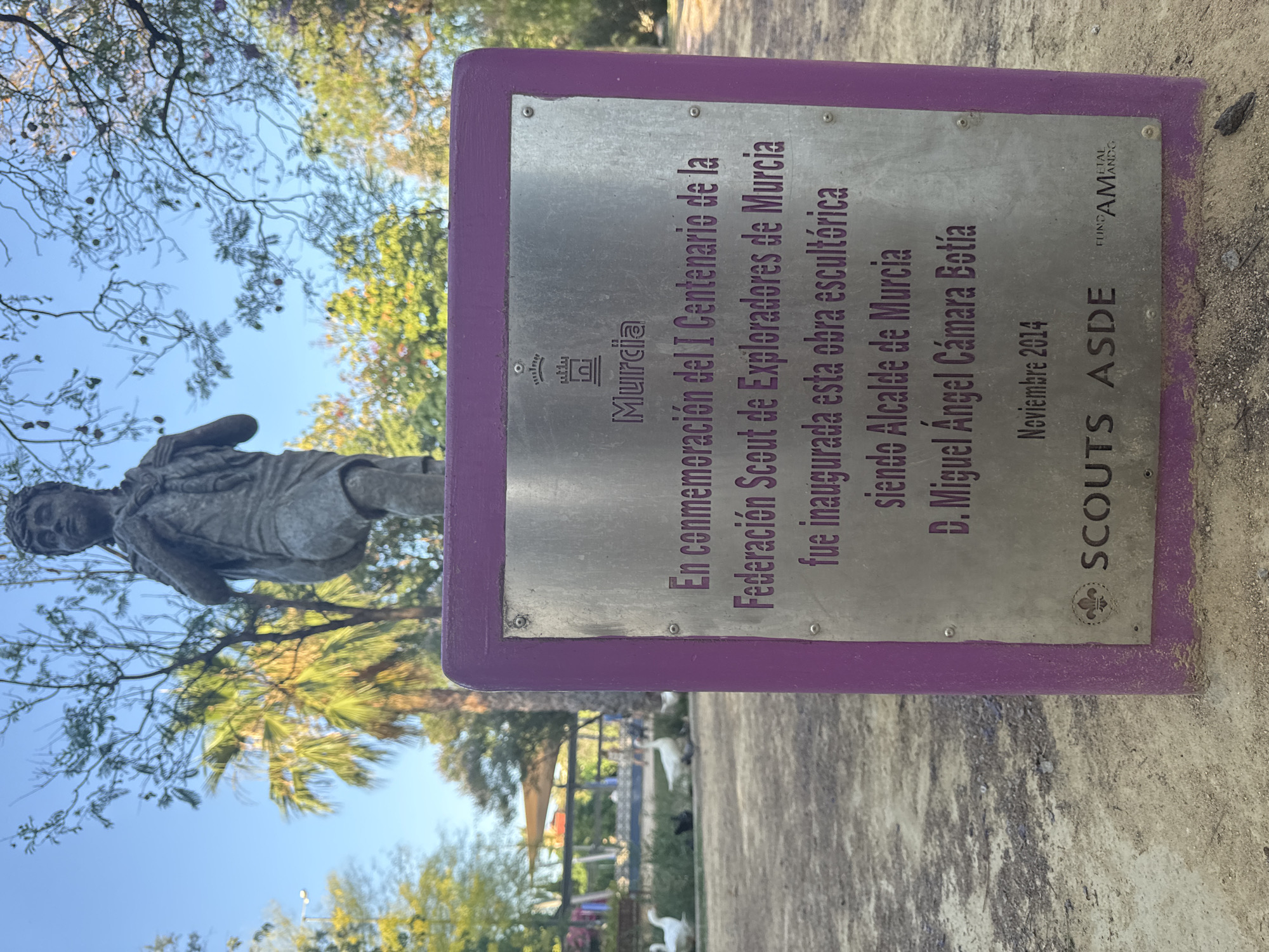 This media file captures a plaque commemorating the I Centenary of the Federation Scout of Explorers of Murcia. The plaque is mounted on a purple frame and made of brushed metal. The text indicates the sculpture was inaugurated when D. Miguel Ángel Cámara Botía was the Mayor of Murcia in November 2014. The plaque also features the logos of Murcia, Scouts ASDE, and FUNDAMANG.
In the background, a bronze statue of a young scout is visible, suggesting the plaque is associated with this sculpture. The statue appears to be in a park setting, with trees and foliage surrounding it. The sunlight suggests it is daytime, and the clear sky hints at fair weather. The scene is set outdoors, likely in a public space in Murcia, Spain, as suggested by the text on the plaque. The overall atmosphere is one of commemoration and celebration of the Scout movement's history.