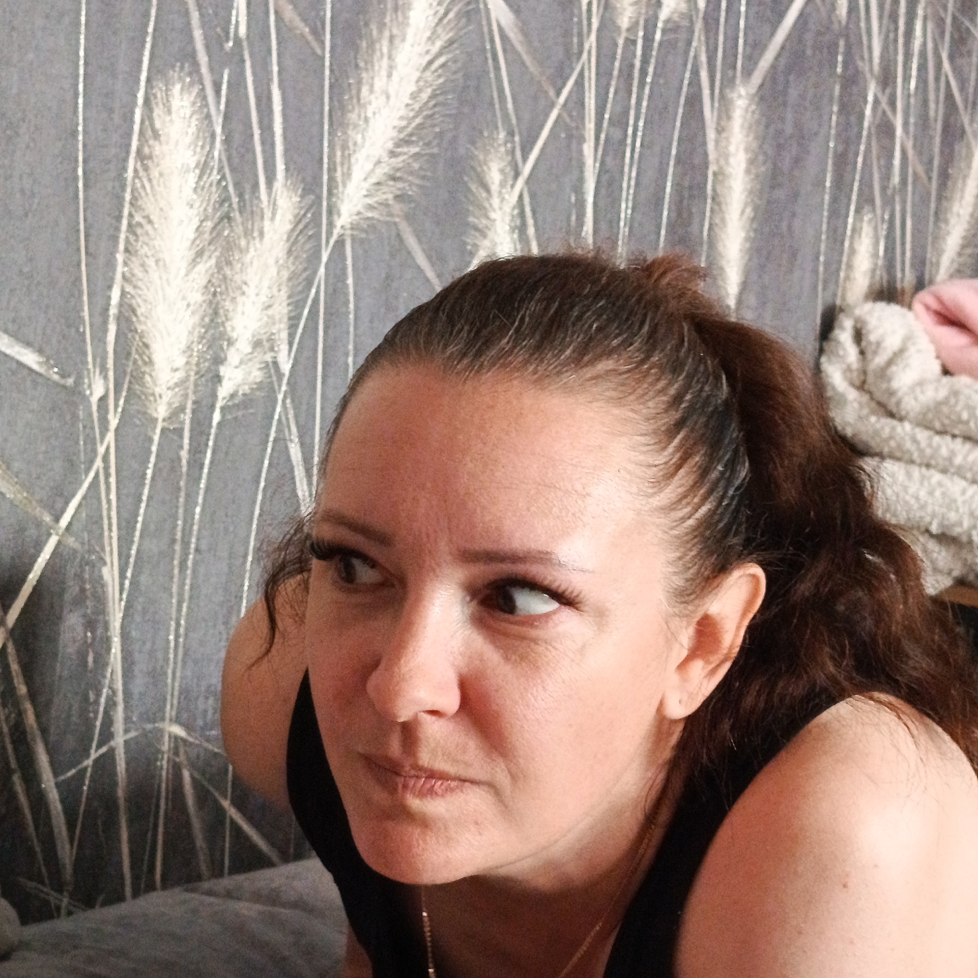 A woman with brown hair pulled back in a ponytail is looking off to the left. She is wearing a black sleeveless top and a gold necklace. The background is a gray wall with a pattern of white, stylized grass or wheat. There is a pile of soft-looking fabric, possibly towels or a blanket, to the right of the woman. The image appears to be taken indoors. There is no information provided about the location within the city of Марковский, Russia.