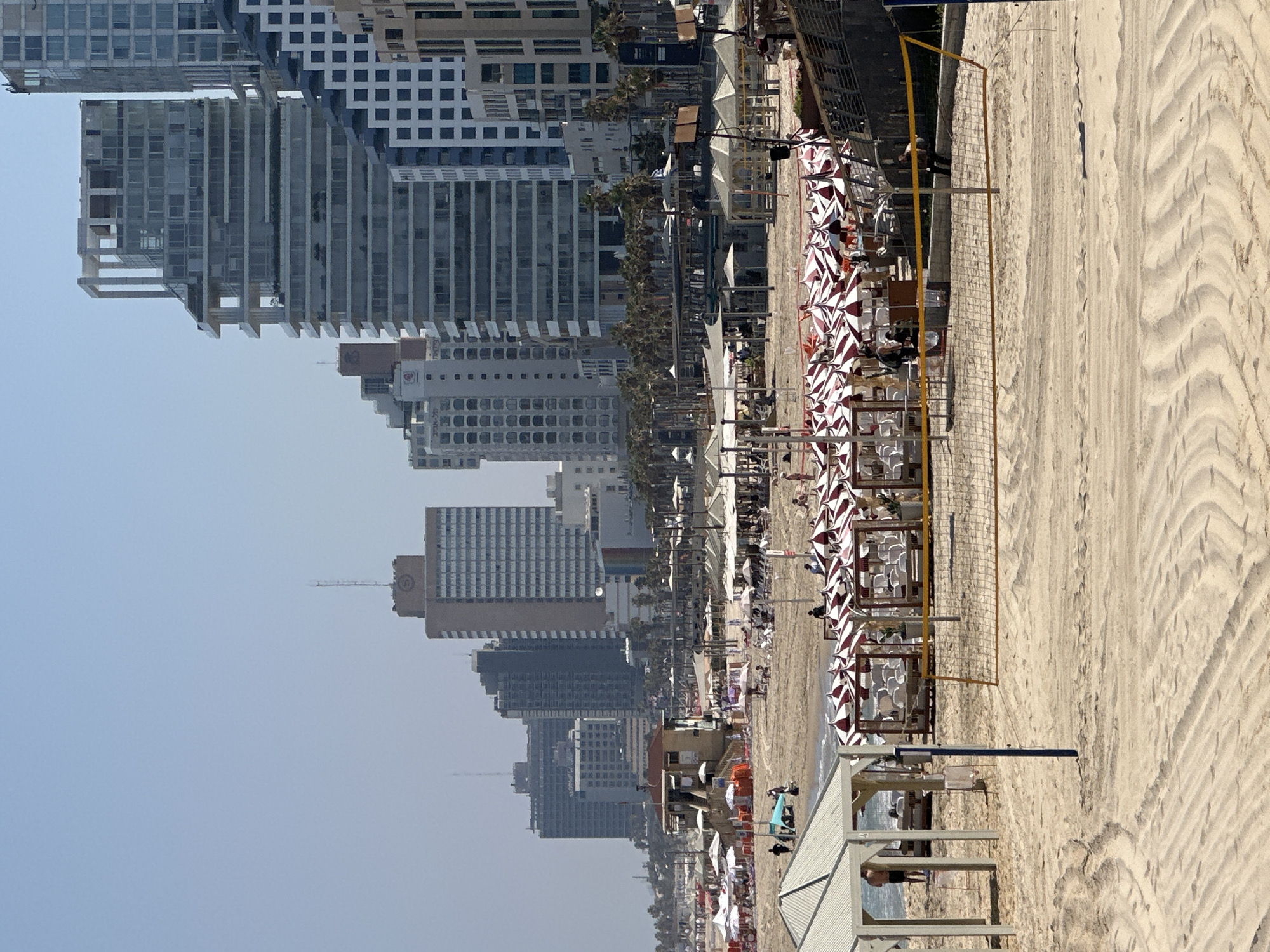 The image shows a sunny day at a beach in Tel Aviv, Israel, with the city skyline in the background. The foreground features a sandy beach with a volleyball net set up. Beyond the net, there are rows of beach chairs and umbrellas, indicating a popular recreational area. In the mid-ground, a lifeguard station stands on elevated stilts, and further back, a line of palm trees separates the beach from the modern city buildings. The buildings in the background are a mix of contemporary high-rises and older structures, reflecting the urban landscape of Tel Aviv. Several people are visible on the beach, some walking, others relaxing under the umbrellas, suggesting a vibrant atmosphere. The sky is clear and blue, typical of a summer day.