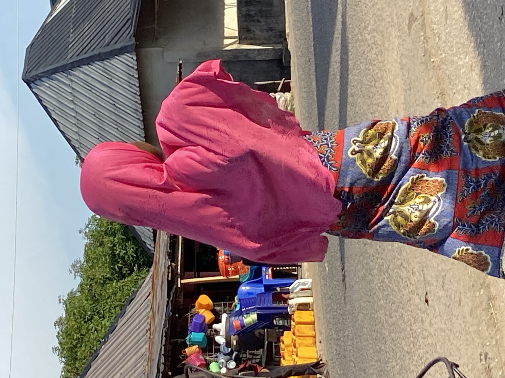 A person wearing a pink head covering and a colorful patterned skirt stands on a paved street in Mararaba, Nigeria. Along the street, there is a shop with colorful plastic items for sale. A corrugated metal roof overhangs the shop. Trees and a bright sky are visible in the background.