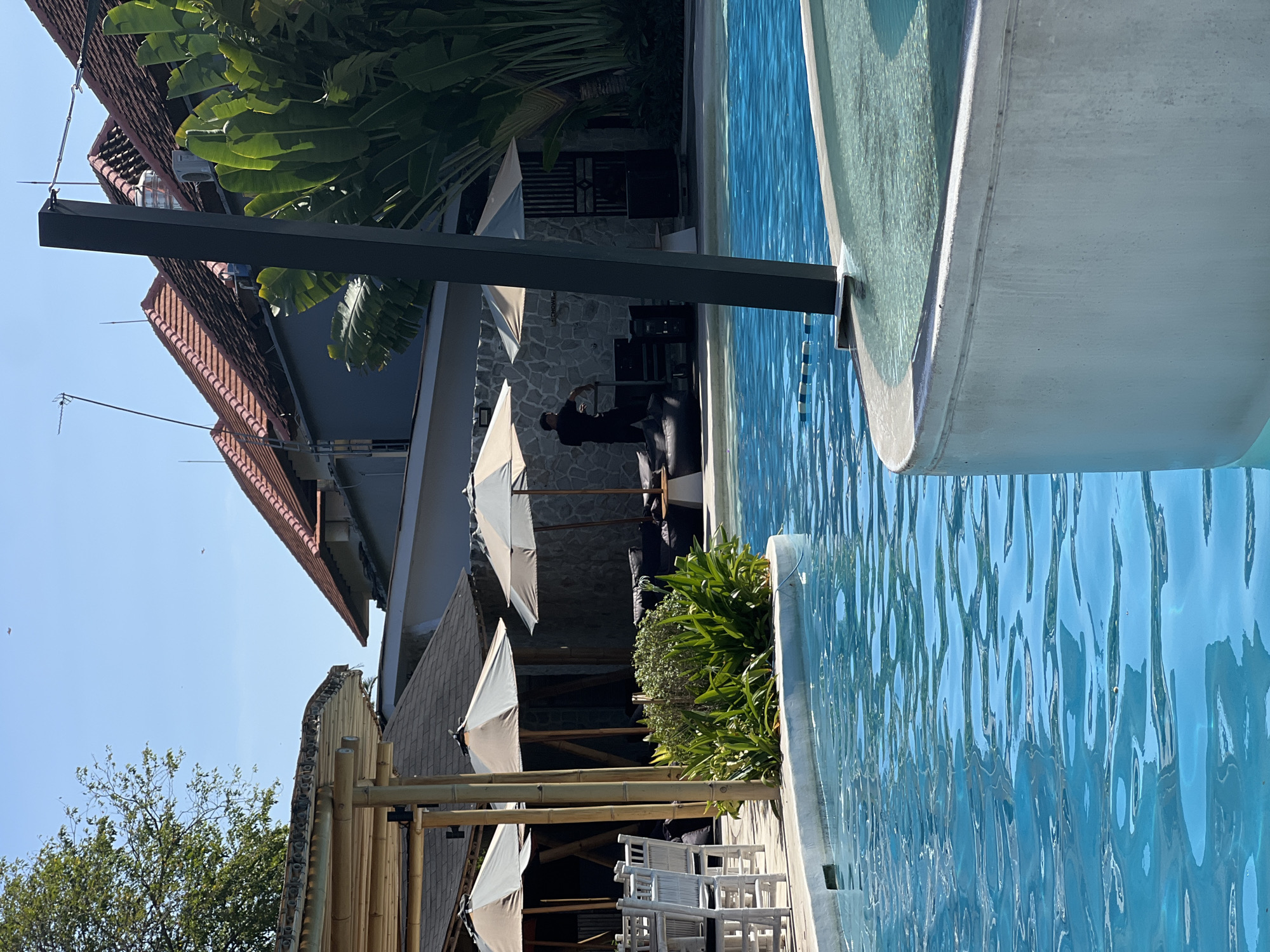 A sunny outdoor scene shows a swimming pool with bright blue water in the foreground. The pool has a curved concrete edge. In the background, there is a covered seating area with tables and chairs, and large leafy plants. A person in a black shirt and pants is standing near some audio equipment. Buildings with red tiled roofs are visible behind the seating area. This appears to be a resort or restaurant in Denpasar, Indonesia.