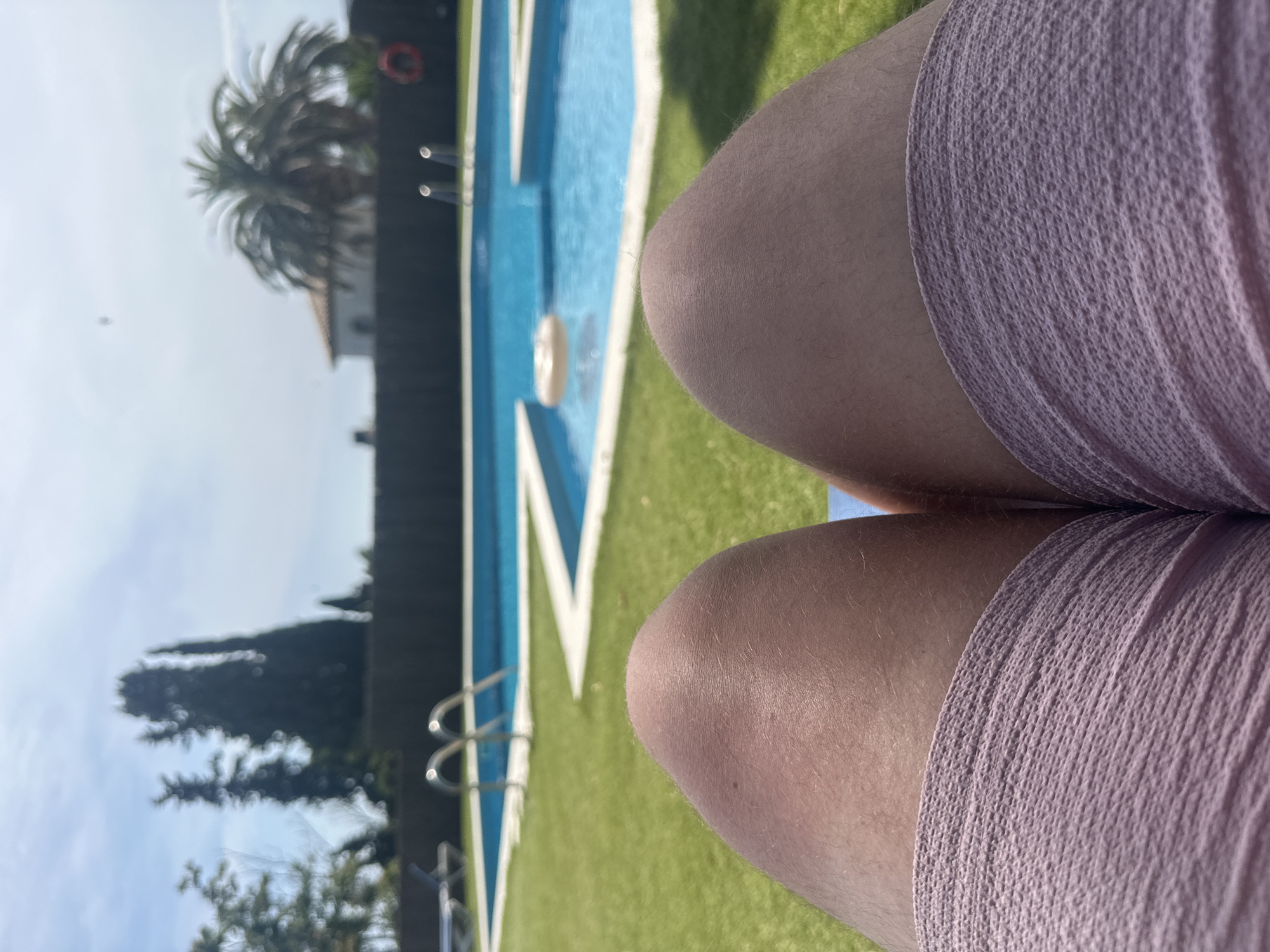 The image depicts a close-up view of a person's legs, possibly resting on a lounge chair in an outdoor setting. The foreground focuses on the individual's thighs and the upper portion of their shorts, which appear to be a light, textured fabric. The background reveals a swimming pool area, complete with blue water, white borders, and a ladder leading into the pool. A light-colored inflatable ring is visible floating in the water. Surrounding the pool is a well-maintained green lawn. In the distance, trees, a palm tree, a building with a red-tiled roof, and a dark fence are discernible. The sky is bright and clear, suggesting a sunny day. The scene is likely situated in a residential or resort area, possibly in a location like Sant Feliu de Guíxols, Spain, known for its coastal resorts and pleasant climate.