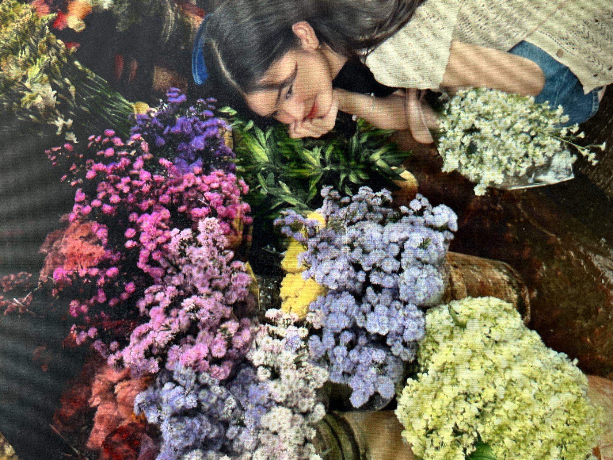 The image shows a young woman with dark hair, wearing a light-colored, crocheted top and blue jeans. She is seated among a vibrant display of flowers. To her left are large bunches of small, clustered flowers in shades of purple, pink, and white. In the foreground, to the lower left, is a large bouquet of pale yellow flowers. To the lower right, another bouquet of small white flowers is visible. The background is filled with more flowers and greenery. The woman is looking to her left, with her chin resting on her hand, and a slight smile on her face. The setting appears to be a flower market. This image was taken in the city of Sejahtera, Indonesia.