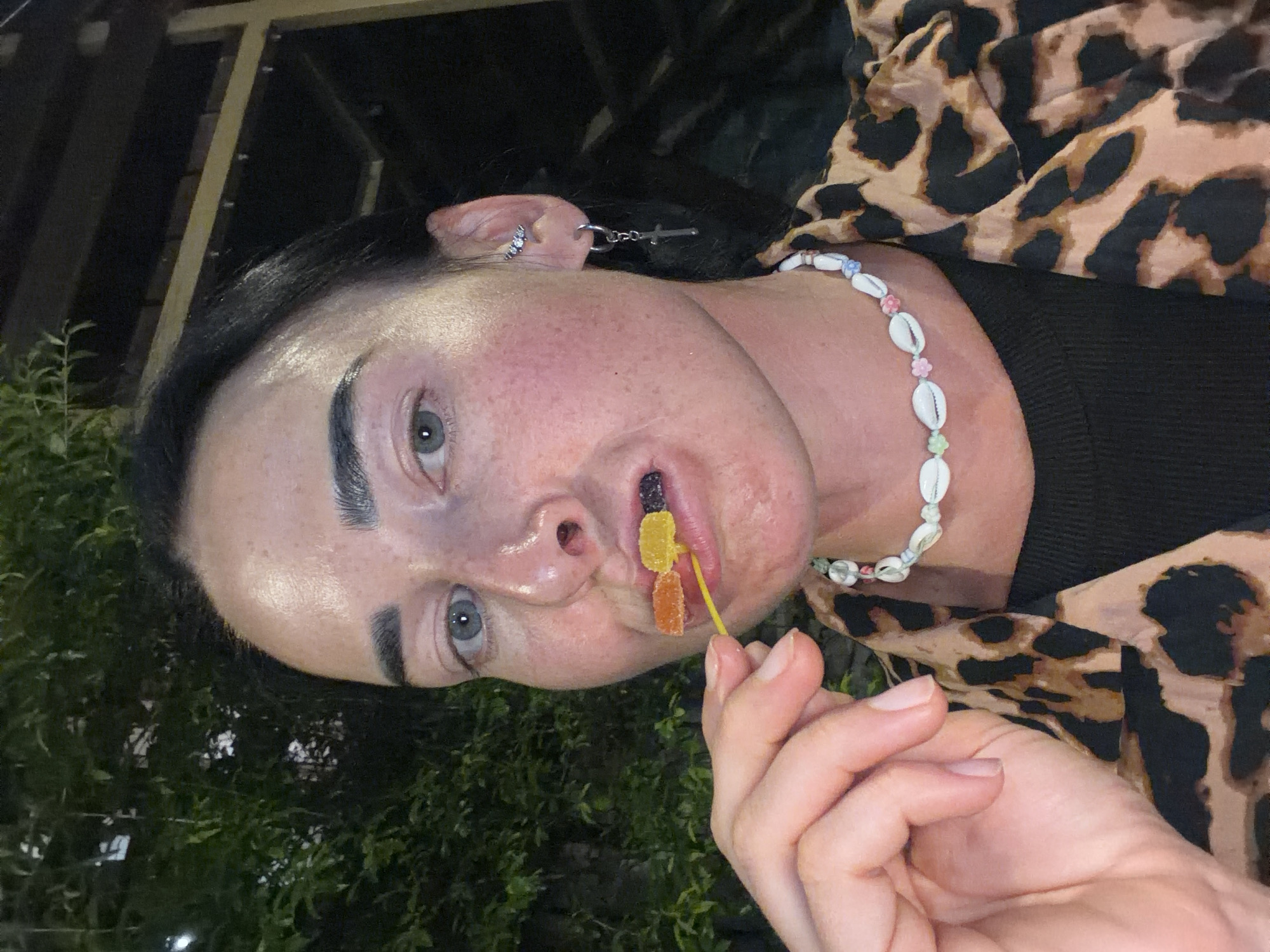 The image shows a close-up of a person holding three gummy candies on a yellow toothpick to their mouth. The person has light blue eyes, dark eyebrows, and is wearing a leopard-print shirt and a shell necklace. The background is dark, with some greenery visible, suggesting an outdoor setting at night. There is no identifiable information about the location being Sochi, Russia.