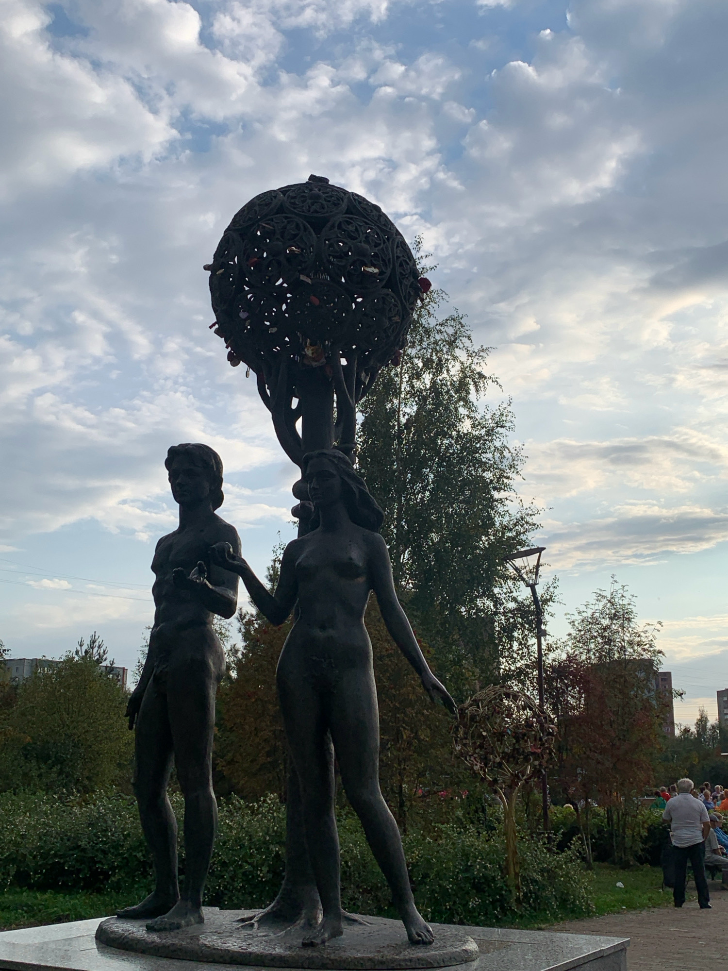 The image shows a bronze statue of Adam and Eve in a park in Prokopyevsk, Russia. Adam stands to the left of Eve, holding an apple. Eve stands to the right, reaching for the apple. Behind them is a stylized tree with a globe-like structure at the top, adorned with numerous small locks. The sky above is partly cloudy, and trees and buildings are visible in the background. Several people are gathered in the park, observing the statue.