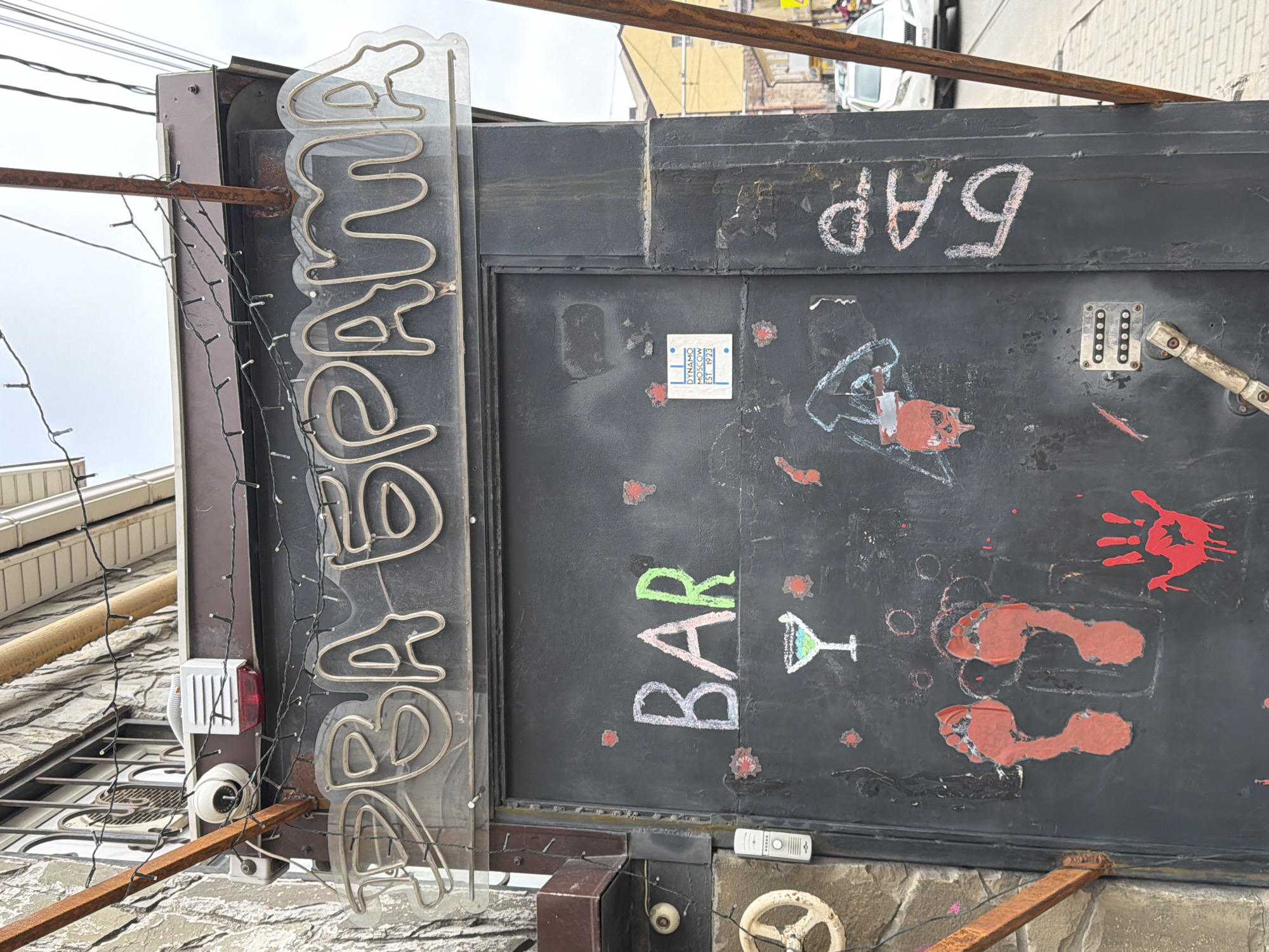 The image depicts the exterior of a bar, possibly in Rostov-on-Don, Russia, based on the neon sign that spells out "ABAANA" in Cyrillic (likely "AVANA"). The black door/signboard is adorned with graffiti and chalk drawings including the word "BAR" and a cocktail glass, as well as a handprint and footprints painted in red. The word "БАР" ("BAR" in Russian) is written in white chalk at the top of the signboard. There is a small blue and white sticker with Cyrillic text ("DINAMO MOSCOW EST. 1923"). A security camera, an alarm system, and various electrical wires are also visible. A rusty metal support structure surrounds the sign. The background shows a narrow alleyway, indicating a possible location in an urban setting.