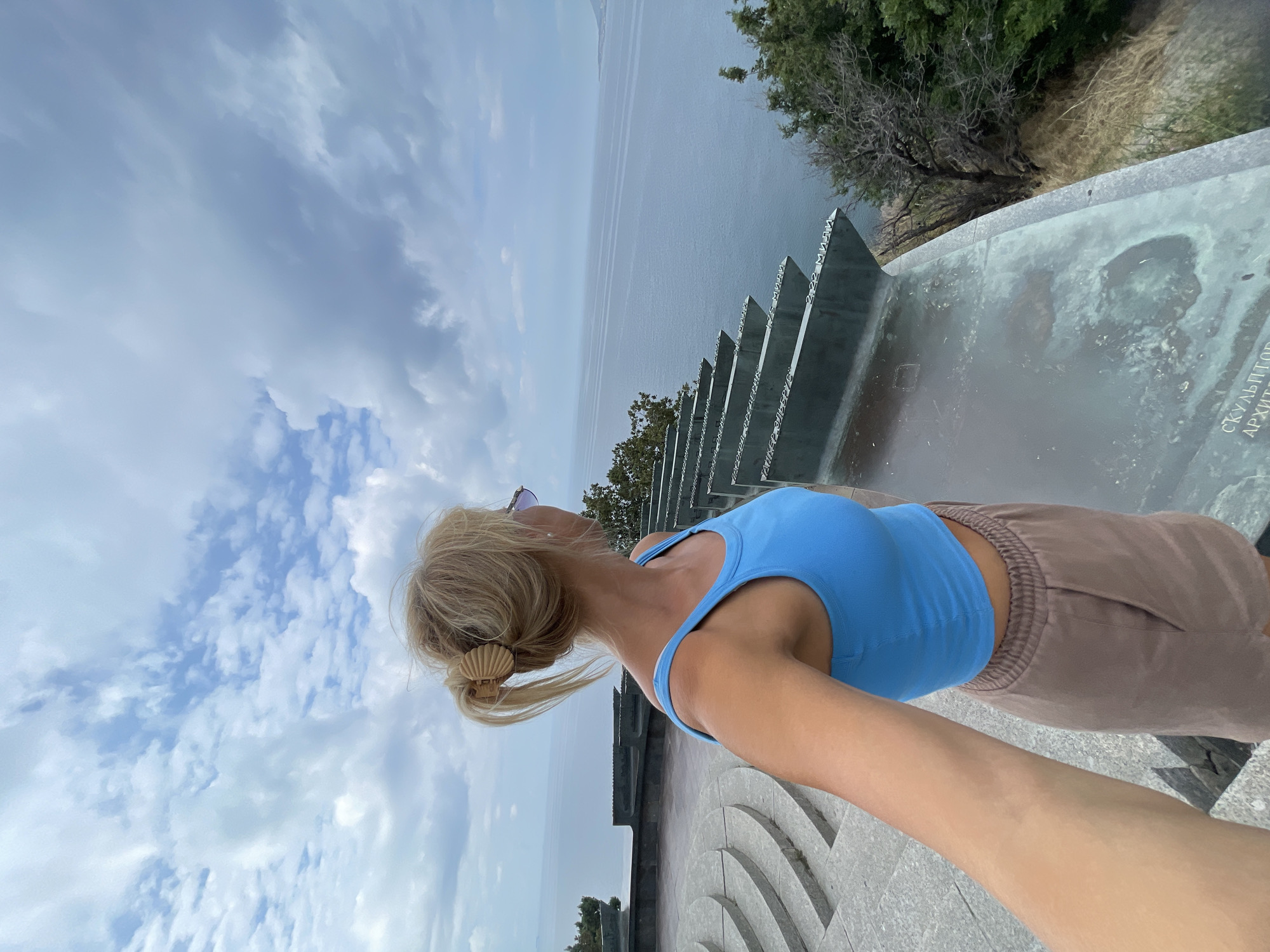 A blonde woman with her hair in a bun held by a clip stands on a stone-paved observation deck overlooking a calm sea under a cloudy sky. She is wearing a bright blue tank top and light brown shorts, and she is taking a selfie. In the background, a long, angled metal structure with inscriptions is visible, as well as some greenery. The observation deck is part of a memorial complex, likely the Malaya Zemlya memorial complex in Novorossiysk, Russia, which commemorates a World War II landing operation. The sea stretches to the horizon, and a distant coastline is faintly visible.