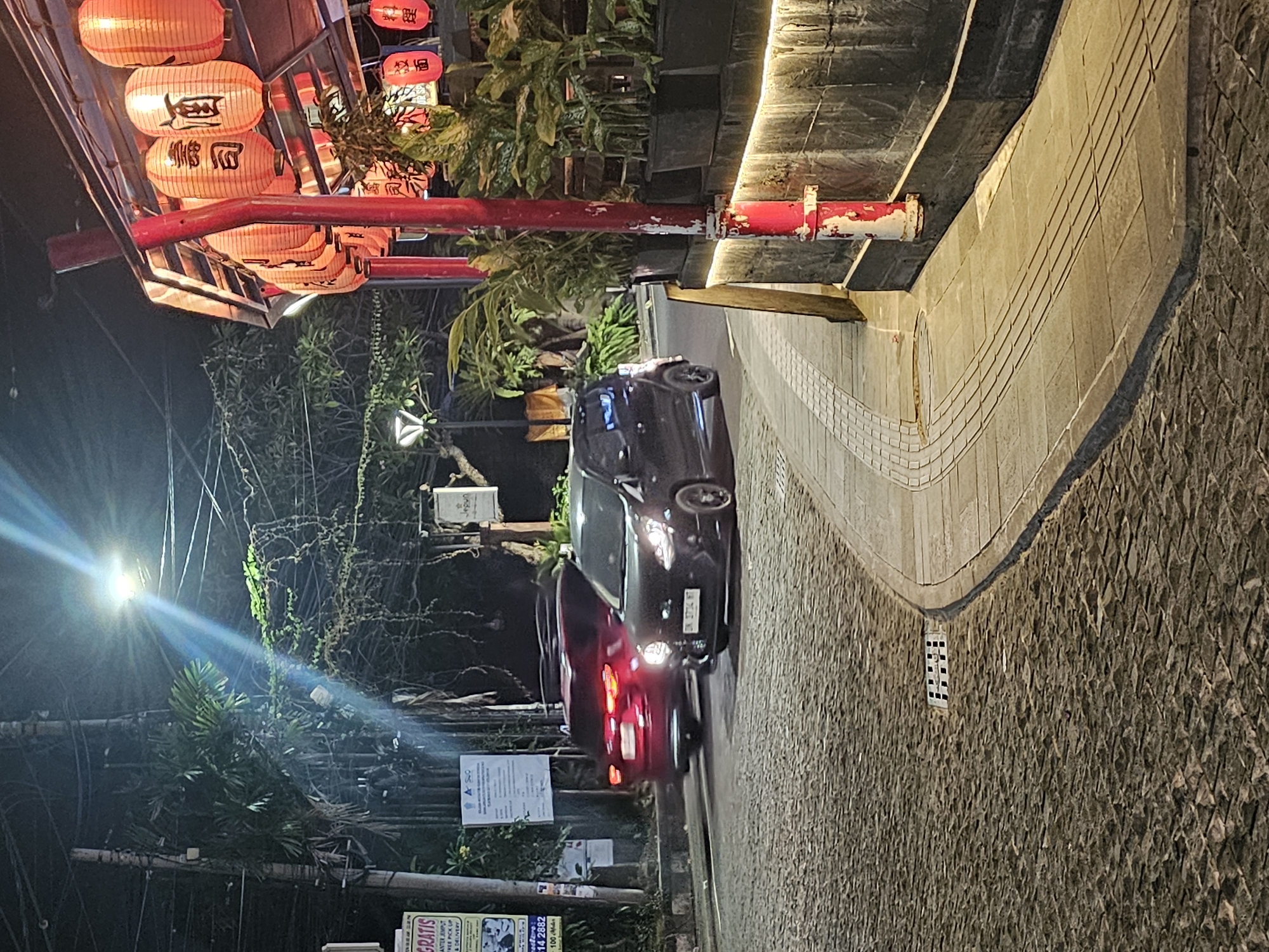 The image shows a street scene at night in Denpasar, Indonesia. Overhead, traditional red Chinese lanterns are strung along a decorative structure. On the left side of the image, street trees and utility lines stretch towards the top of the image where a street light shines, creating a lens flare. Along the right side is a walkway made of patterned stone, with an adjacent wall also constructed of rough-cut stone.
A dark-colored car is driving on the street between the Chinese lanterns and the walkway. Its headlights and taillights are illuminated. In the lower left, there are street signs, including one that says "GRATIS ANTER JEMPUT FREE PICK UP & DELIVERY" with a phone number below. Above this sign is another sign containing a considerable amount of text. The car's license plate indicates the car is from Indonesia.
