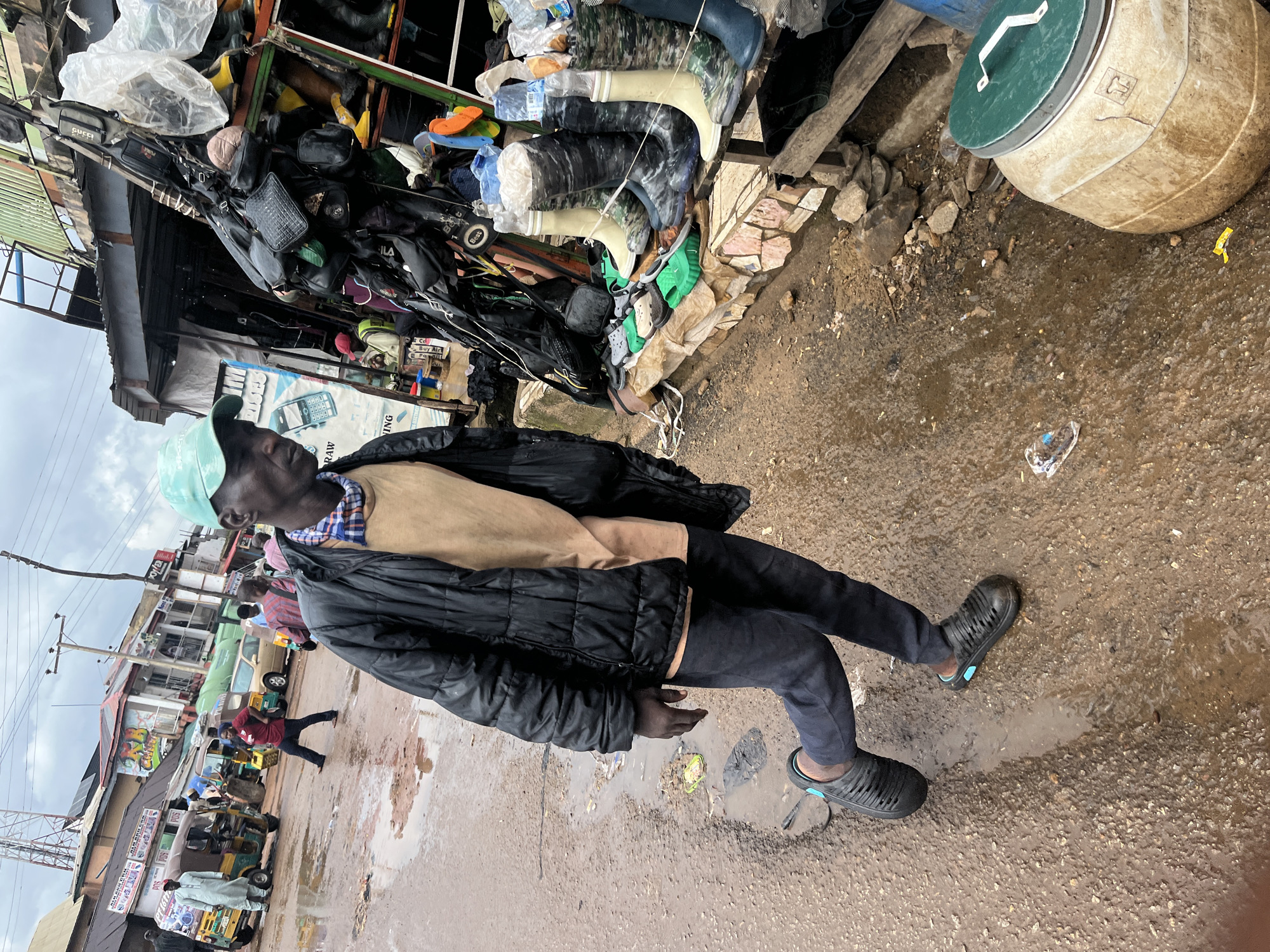 This is a vertical, medium-shot, eye-level photograph of a busy street market in Jos, Nigeria, under an overcast sky. The foreground is dominated by a man in a black puffer jacket, a light brown t-shirt, and dark pants, wearing black slip-on shoes with blue accents. He is walking towards the viewer's left, with his body angled slightly away. His face is visible, and he wears a light blue baseball cap.
The man is positioned in front of a stall that displays a variety of footwear, primarily boots and sandals, arranged on shelves. The stall is packed with goods, including bags and other items hanging from above. The ground is muddy and wet, with puddles reflecting the cloudy sky, indicating recent rain.
In the midground and background, the market continues with more stalls, a few vehicles including auto-rickshaws (popularly known as Keke Napep), and other people moving about. Buildings line the street, some with colorful signs and awnings. The overall atmosphere suggests an active commercial area. A large, beige plastic barrel with a green lid is visible in the lower right corner of the image.