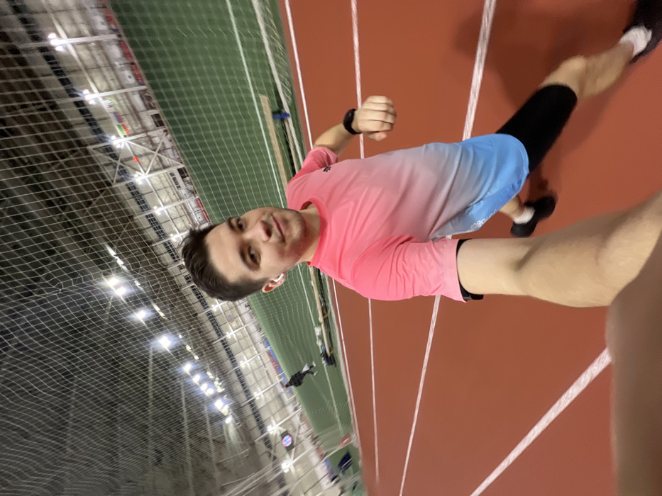A man poses for a selfie on an indoor track. He wears a pink and blue shirt, black compression socks, and running shoes. The track is red with white lane markers. A green netted area with stadium seating is visible in the background. The building has a high ceiling with industrial lighting. The man is running mid stride.