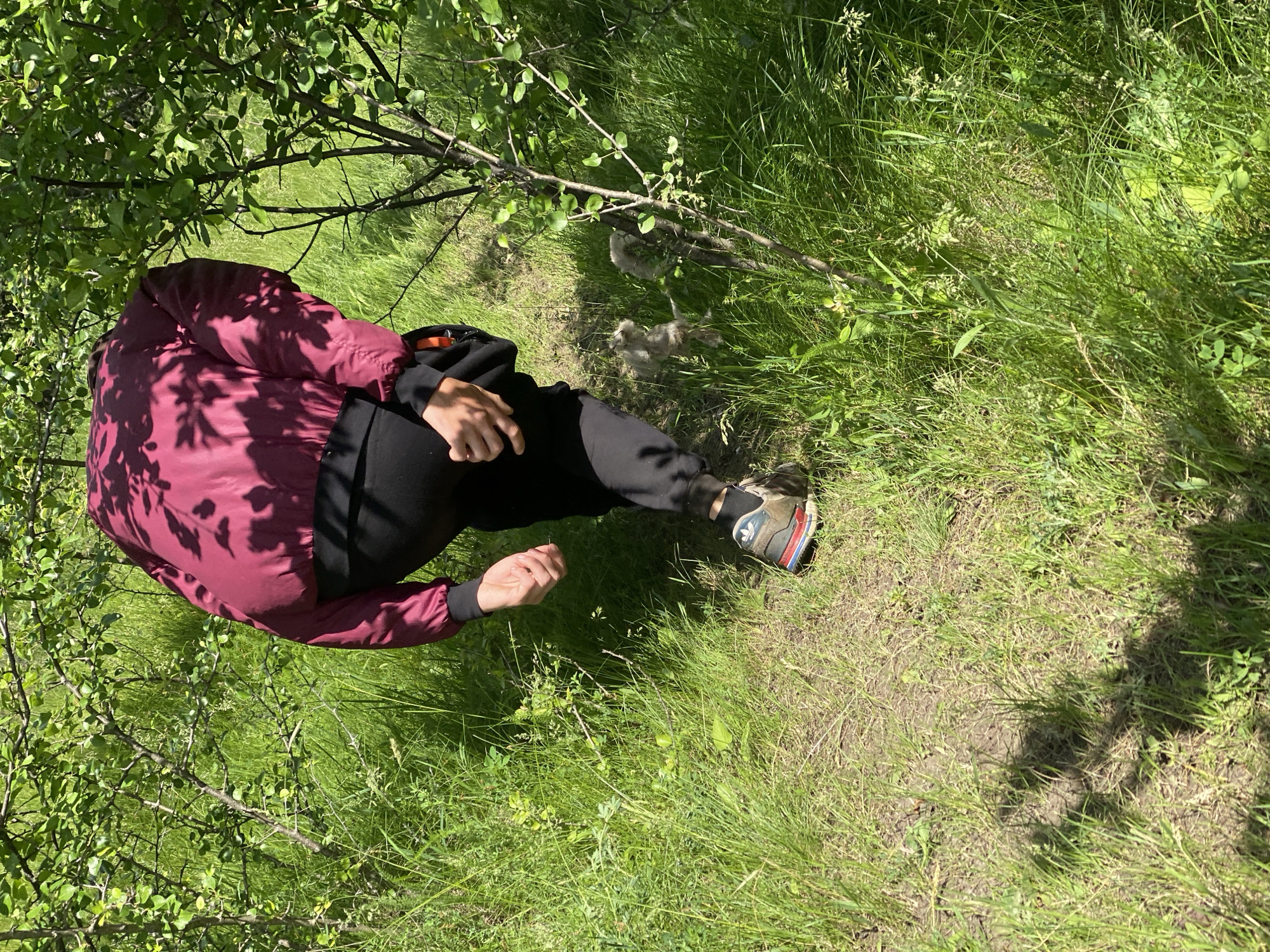 A person wearing a dark red jacket, black pants, and a hiking shoe is crouching in a grassy area. The person is near some sparse brush and tall grass, potentially in a wooded area. The location is the city of Ustyvytsia, Ukraine.