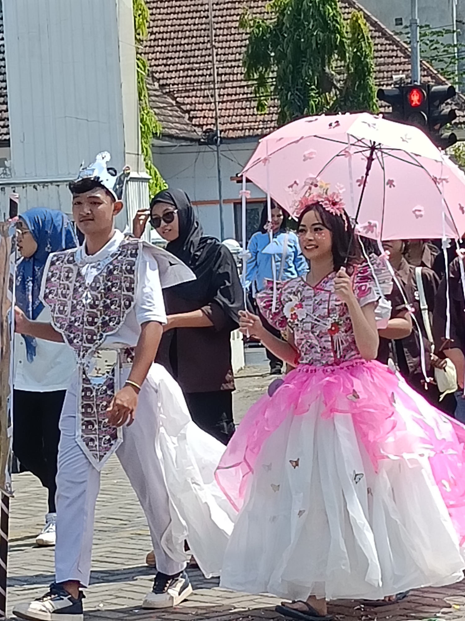In Kediri City, Indonesia, a festive parade is underway. A young male, adorned in a white outfit and a silver crown, walks alongside a young female dressed in a pink and white layered gown. She holds a pink umbrella decorated with floral patterns. Both are part of a procession on a paved street, with buildings and trees visible in the background. A traffic light, showing a red signal, is positioned on the right side of the image. The atmosphere appears to be celebratory, with participants dressed in elaborate costumes.
