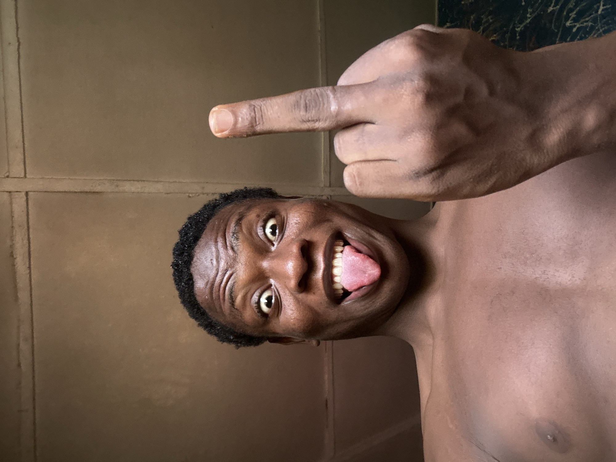 The image shows a dark-skinned male, shirtless, displaying his middle finger with his tongue out. The background is a beige wall with a visible seam and a dark, patterned surface in the upper right corner. The location is unknown.