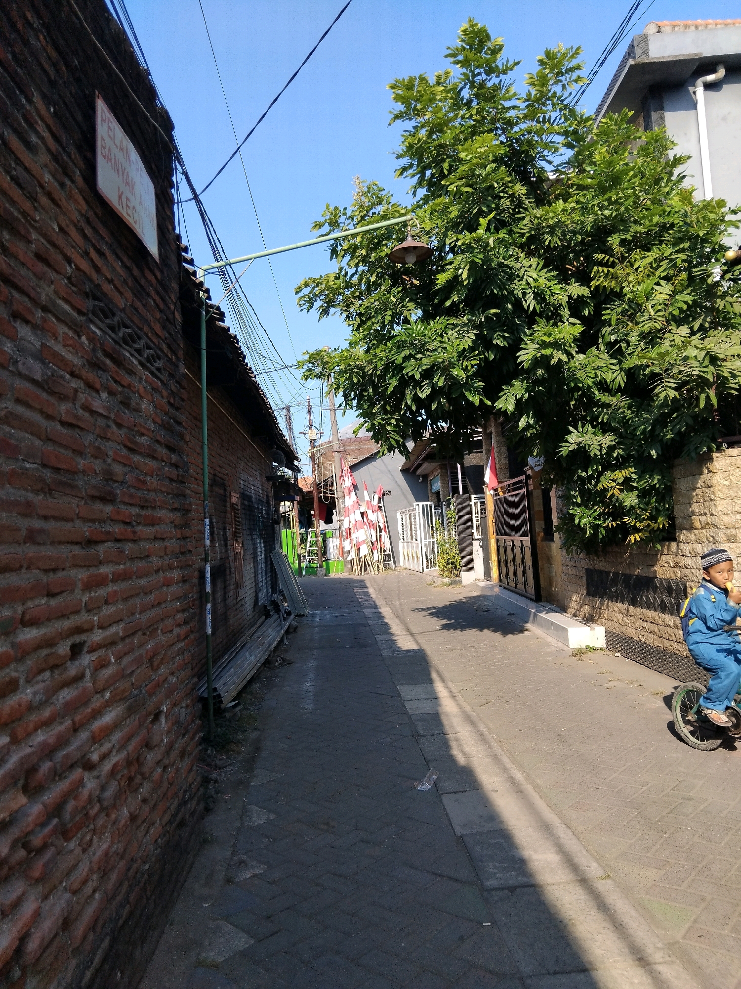 The image depicts a narrow, paved alleyway in the city of Kepuh Kiriman, Indonesia, under a clear blue sky. The alley is flanked by a weathered brick wall on the left and buildings and a large, leafy tree on the right. A young boy, dressed in a blue uniform and cap, is riding a small bicycle on the right side of the alley, holding what appears to be a popsicle.
The brick wall on the left has a sign with text that is partially legible, appearing to read "PELAN-PELAN BANYAK ANAK KEC". Wires are strung across the alley from poles and buildings. In the distance, red and white flags, possibly Indonesian flags, are visible, suggesting a local celebration or event. There are also stacks of building materials and a ladder leaning against a structure along the alley. The ground is paved with interlocking bricks. Shadows are cast across the alley, indicating the direction of sunlight.