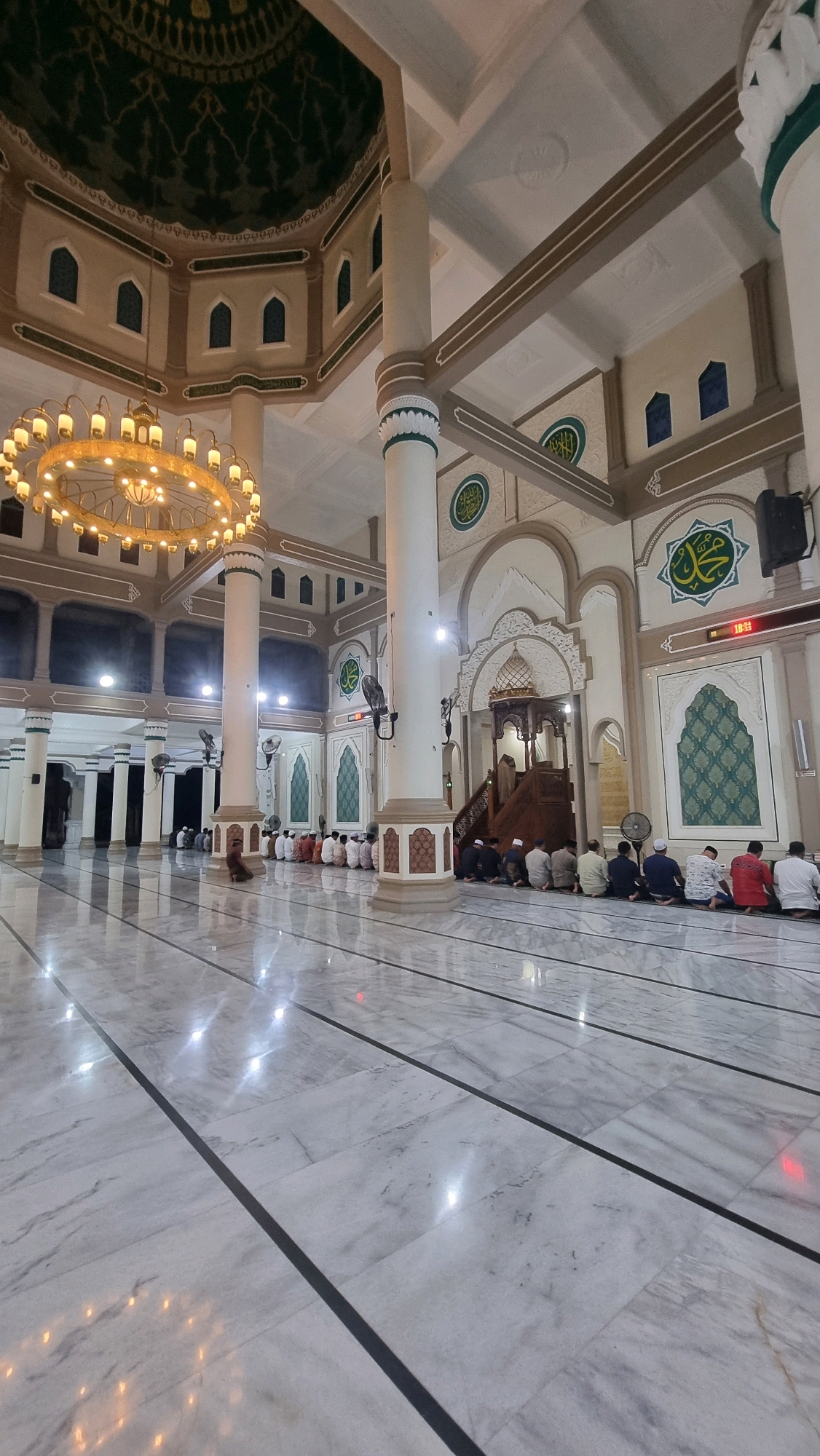 A vertical, eye-level, wide-angle shot captures the interior of a mosque in Lhokseumawe, Indonesia. The scene is dominated by the pristine, white marble floor, which reflects the ornate details of the mosque's architecture. Multiple groups of men, dressed in Islamic attire, are seated on the floor, facing the front of the mosque, indicating they are engaged in prayer or religious observance. A grand chandelier hangs from the ceiling, casting a warm glow, while intricate patterns adorn the dome and walls. The mosque features tall, cream-colored pillars and decorative Islamic calligraphy. The overall ambiance is one of solemnity and devotion.