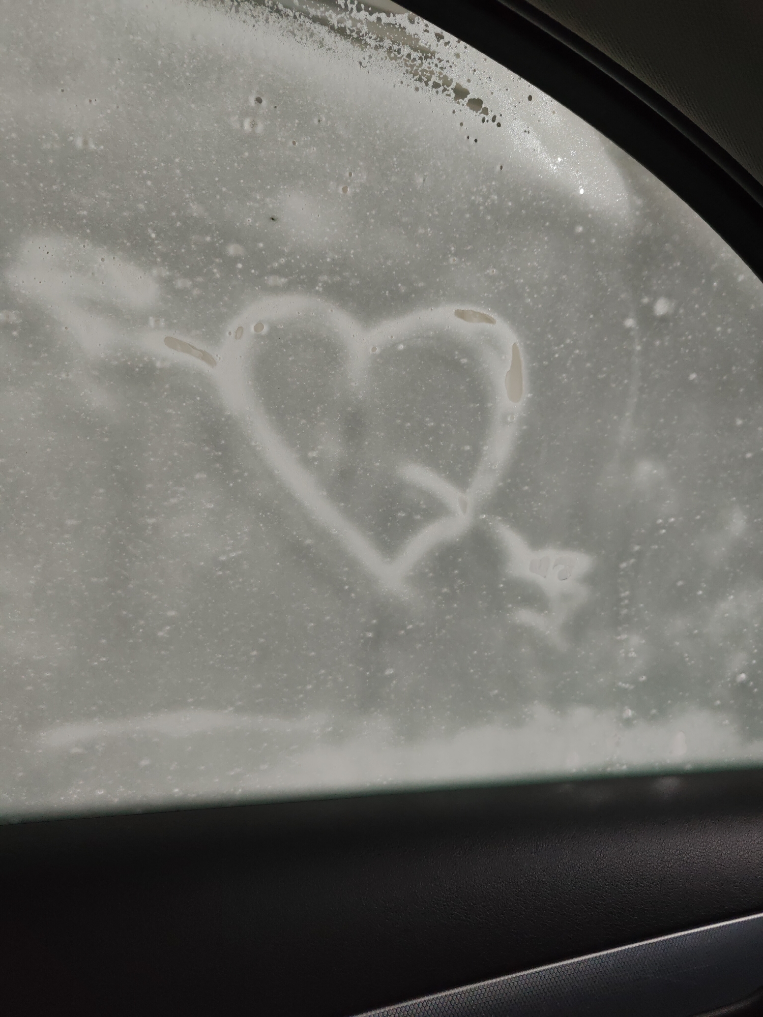 A drawing of a heart pierced by an arrow is etched into a frosted car window. The drawing is created by wiping away the frost, revealing the glass underneath. The heart is symmetrical with a slight point at the bottom. The arrow passes through the center of the heart and has fletching at the tail end. The background is a blurry expanse of white frost with small water droplets scattered across the surface. The lower portion of the image shows a dark, textured surface, likely the interior of a car, with a thin silver trim. The overall impression is one of a cold, possibly winter day, given the frosted window. The drawing suggests a romantic or sentimental gesture. No text is visible in the image. The location is Chaikovsky, Russia.