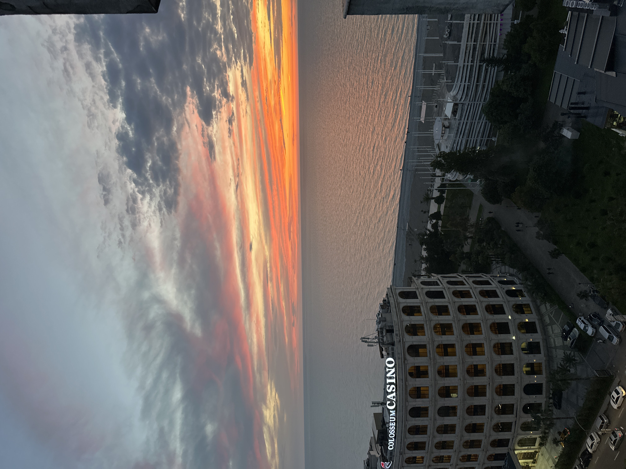 The image depicts a sunset view in Batumi, Georgia. The sky is filled with gray clouds with streaks of orange and yellow near the horizon. Below the sky is the sea, reflecting the colors of the sunset. On the right side of the image, there's a portion of the city. The Colosseum Casino is a prominent building, with its name illuminated in white lights. Beyond the casino, there is a street with cars parked and a walkway with trees. The image is taken from a high vantage point, possibly a building or an elevated structure. The scene appears to be captured during the early evening, given the diminishing daylight.