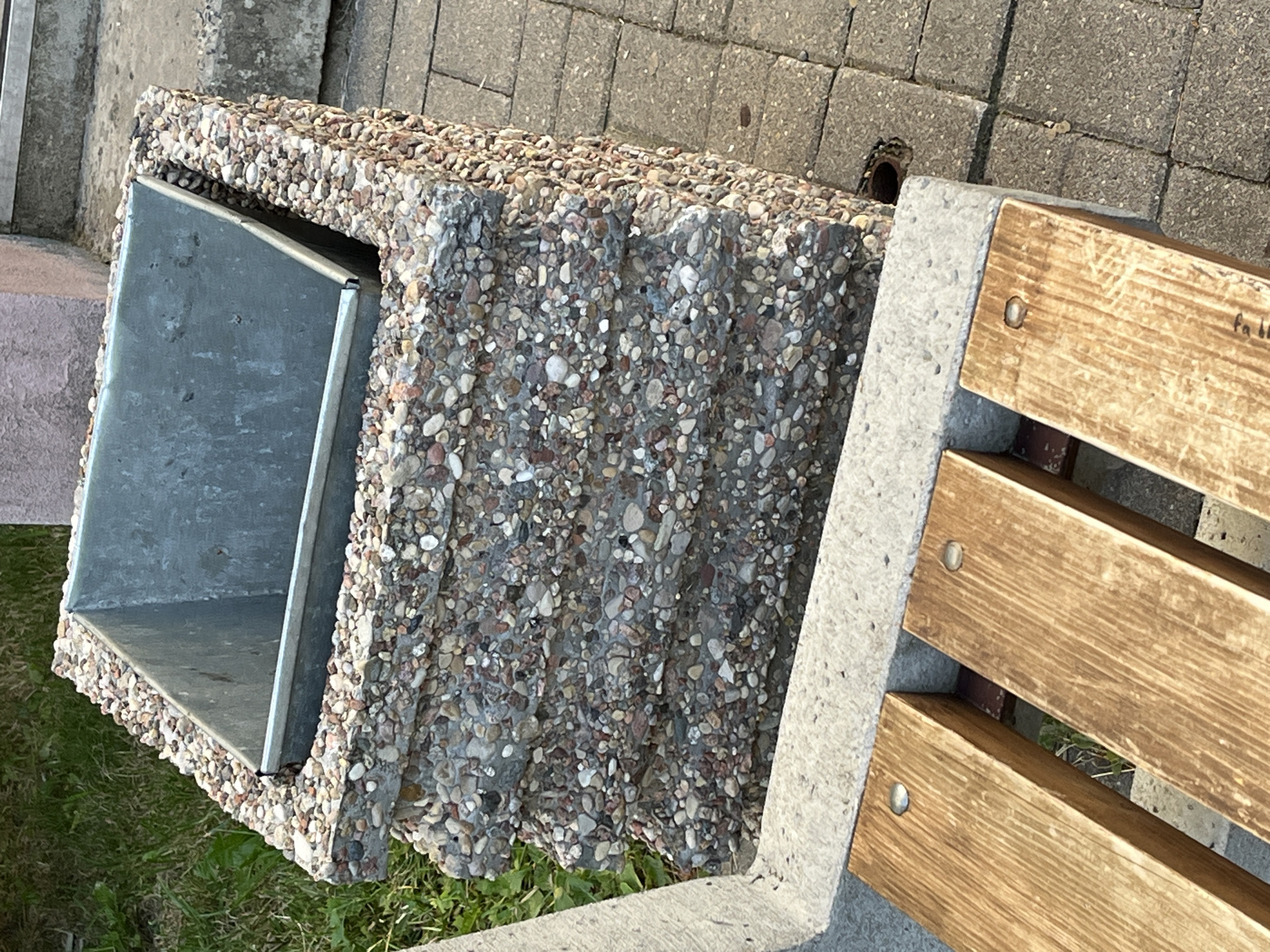 The image shows a close-up, eye-level shot of a concrete planter with a metal insert, likely containing soil. The planter is textured with embedded pebbles of various sizes and colors, and it has a tiered, stepped design. To the left of the planter is a portion of a concrete bench with wooden slats, secured by metal screws. The bench appears to be part of a larger seating area. The background includes green grass to the left and paved ground, made of interlocking grey bricks, to the right. A circular drain grate is visible in the pavement. The scene is outdoors, under natural light, suggesting it is daytime. There are no people or animals present in the image. The overall impression is of an urban or park setting. The presence of a planter and bench suggests a public outdoor space. Without any identifiable landmarks or specific architectural styles, pinpointing the exact location within Minsk, Belarus is not possible from the visual information alone.