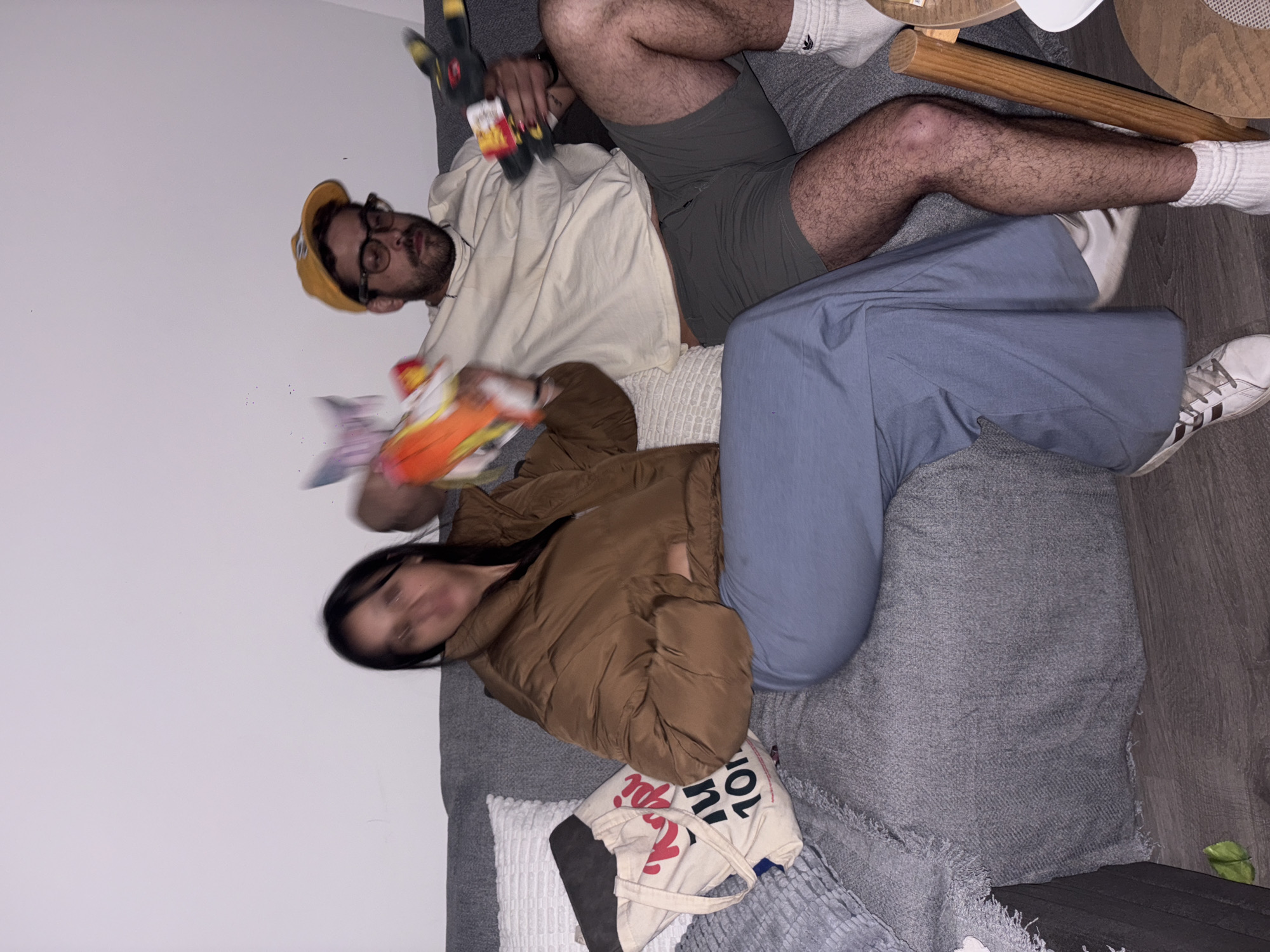 Two people are lounging on a grey sofa in an interior space with a light grey wall on the left and a grey wood floor on the right. One person, wearing a yellow cap, glasses, and a light-colored shirt, is holding a plush toy and a box of crackers. They are positioned partially on top of the other person, who is wearing a brown jacket and blue pants. A tote bag with red and black writing is next to the second person. The location is unknown, and there is no evidence of it being in Mexico City, Mexico. A small wooden table is partially visible in the upper right corner.