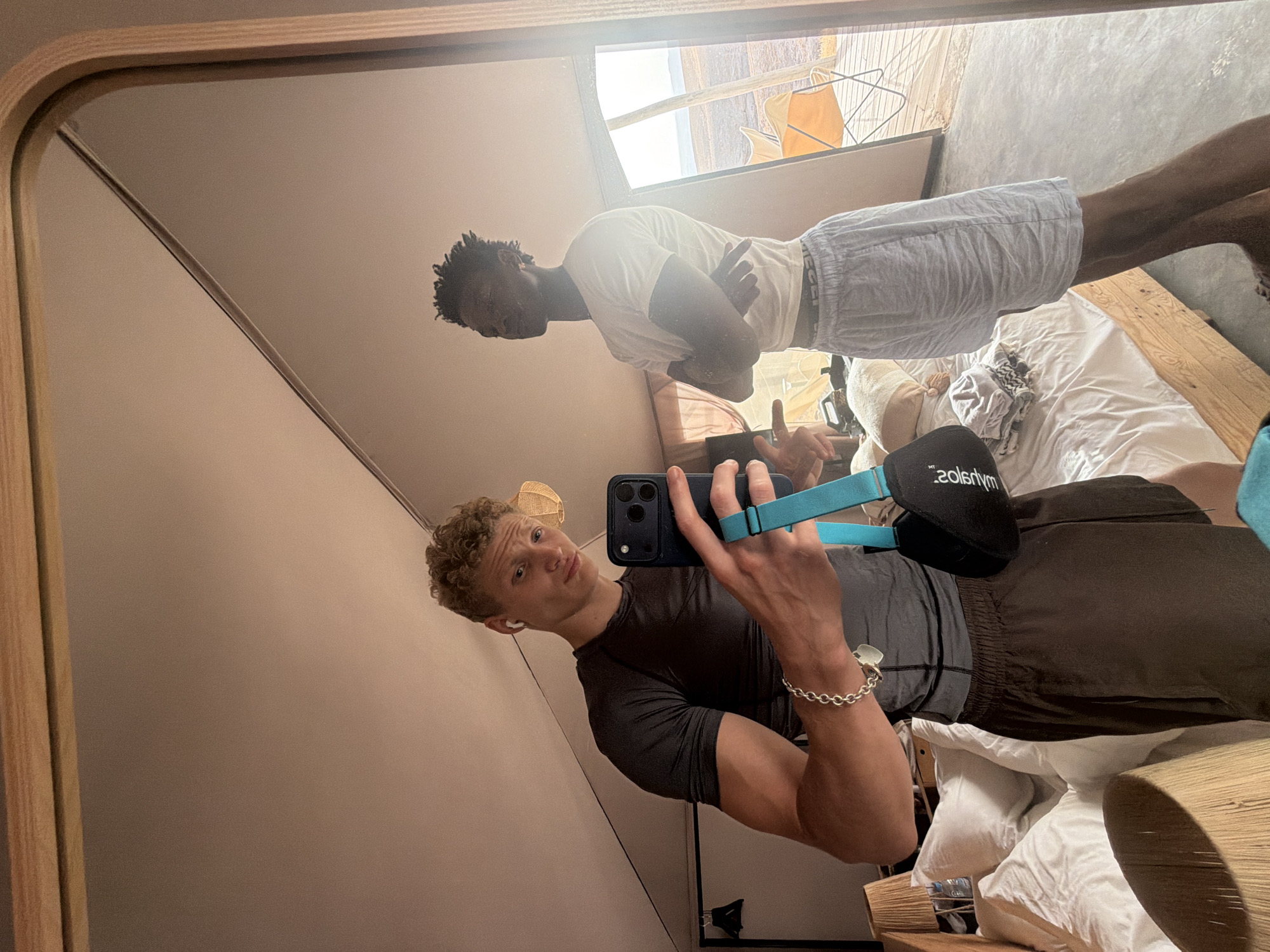 In a tent in Agafay, Morocco, two young men are reflected in a large mirror. The man on the left, with curly blonde hair and wearing earbuds, is taking a selfie with a smartphone. He has a serious expression and is wearing a dark grey t-shirt and brown shorts. He has a silver bracelet on his left wrist. The man on the right, with short dark hair, is looking away with his arms crossed. He is wearing a white t-shirt and light grey shorts. The tent has a wooden bed with white pillows and blankets. In the background, through an open tent flap, a desert landscape with chairs and a blue sky is visible. The scene is lit by natural light, possibly from the setting sun, casting a warm glow.