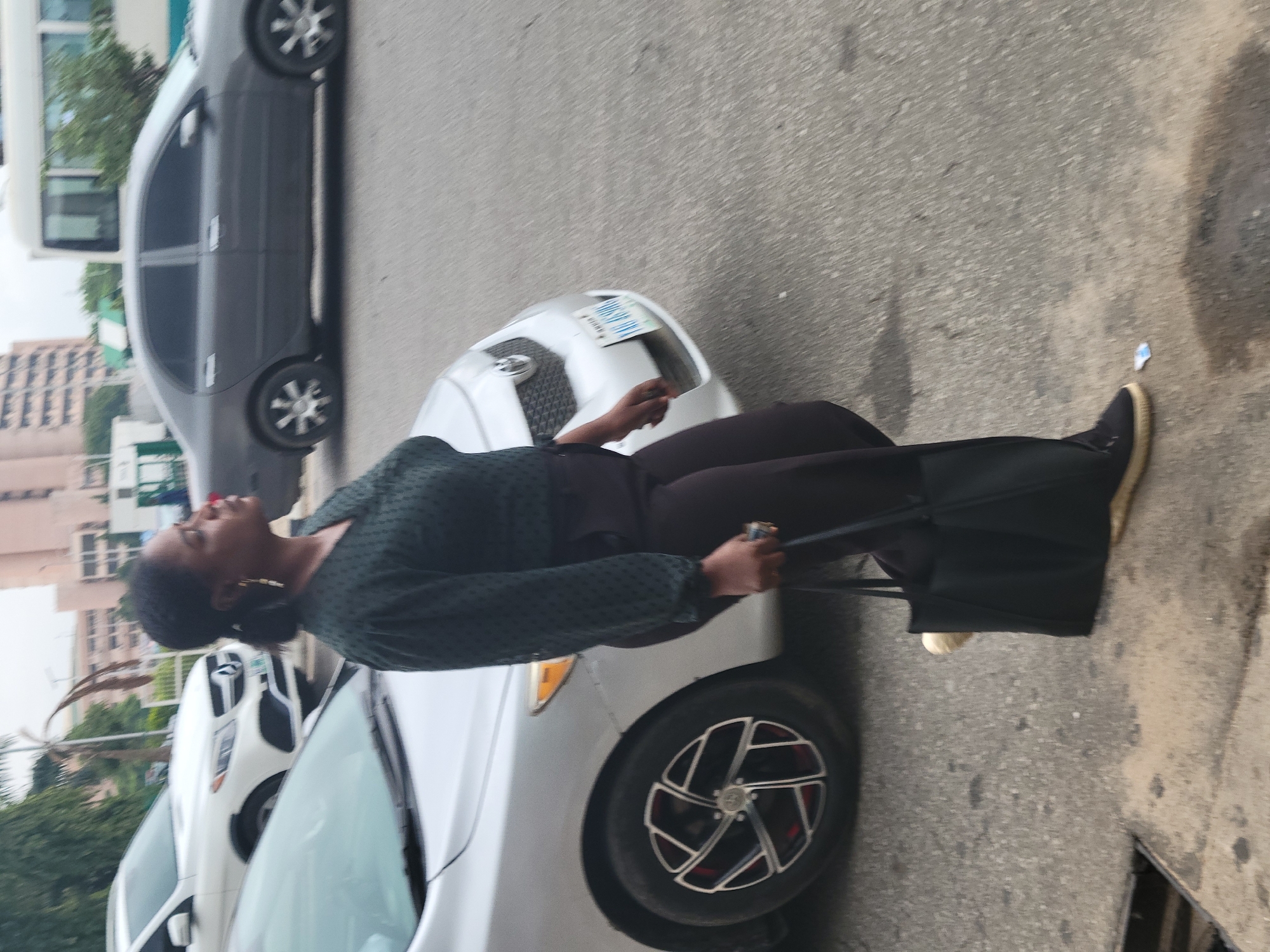 A woman with dark hair, wearing a dark green blouse with a textured pattern and dark pants, is walking on a paved road in Abuja, Nigeria. She is carrying a large black tote bag in her right hand and has a small, light-colored object on the ground near her left foot. The woman is facing to the right and appears to be looking upwards. In the background, a silver sedan is parked on her left, and a dark gray sedan is in the mid-ground. Further in the background, there are buildings, trees, and a white bus. The lighting suggests it is daytime.