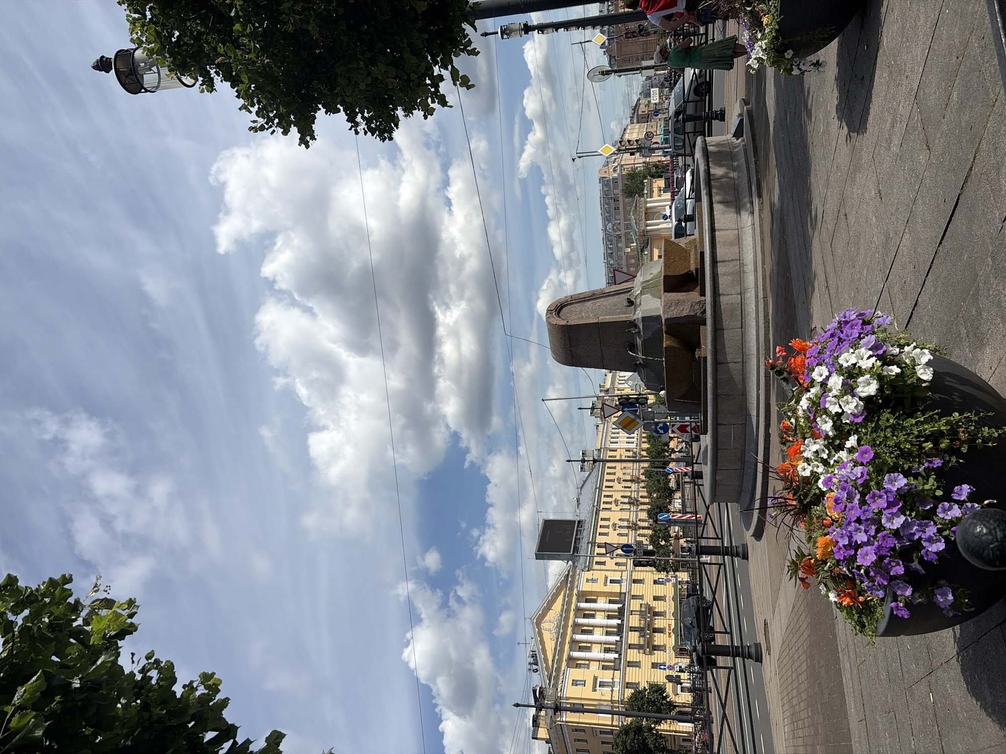 The image depicts a street scene in Saint Petersburg, Russia, likely on a sunny day given the bright lighting and scattered clouds. In the foreground, a large planter box overflows with vibrant purple, white, and orange flowers, adding a splash of color to the paved walkway. To the left, a stately yellow building with classical architectural features, including columns and arched windows, dominates the background. Across the street, a fountain with stone carvings is visible, surrounded by pedestrian areas and a roadway with traffic. Several cars are present on the road, indicating an active urban environment. Trees line the sidewalks, and streetlights stand tall, contributing to the city's infrastructure. The sky above is a mix of blue and white clouds, suggesting fair weather. The overall impression is one of a well-maintained public space in a historic European city.