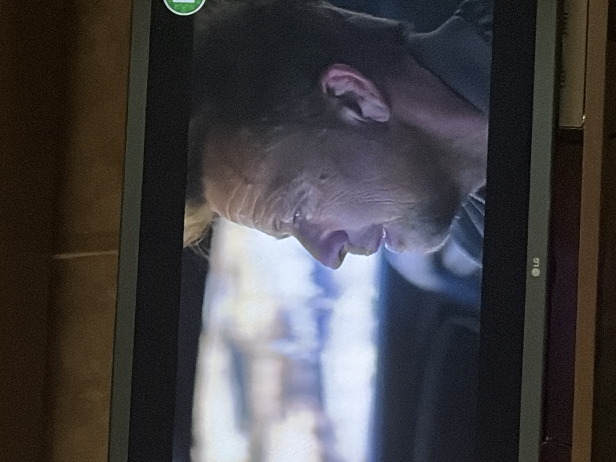 The image is a medium shot of a man's profile from the nose to the ear, shown on a television screen. The man appears to be middle-aged with wrinkles on his forehead and his hair is dark and short. He is wearing a dark shirt. The background is blurred, suggesting he is in a vehicle. The television is placed on a wooden surface. A green circular logo is visible on the right side of the screen. The LG logo is visible at the bottom of the screen.