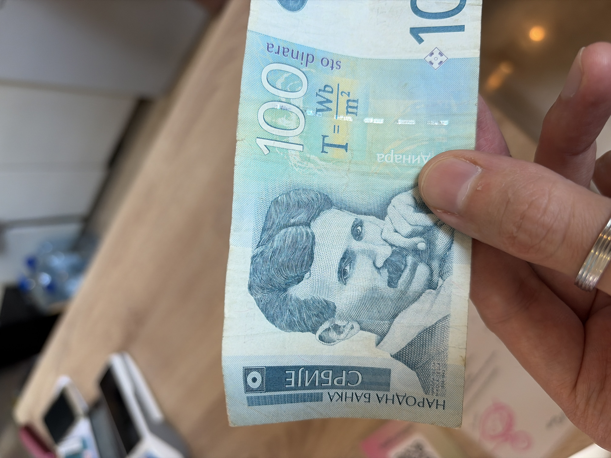 A hand holds a 100 Serbian dinar banknote featuring Nikola Tesla. The banknote has a blue and white color scheme. The scene is indoors, with a wooden table visible in the background. A cash register is also visible in the background. No specific information to locate the scene in the city of Budva, Montenegro.