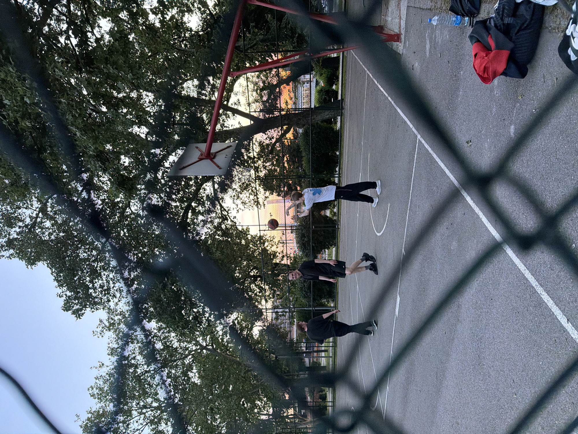 A basketball court is seen through a chain-link fence. Three people are present on the court. One is wearing a white shirt and black pants, and is positioned with a basketball in the air, seemingly in the act of shooting. Two other individuals are wearing black clothing. Clothing and a water bottle rest along the sidelines. The backdrop of the court shows trees and the skyline of Belgrade, Serbia. The image is taken at dusk, indicated by the sky. A basketball hoop and backboard are positioned on the left side of the frame.