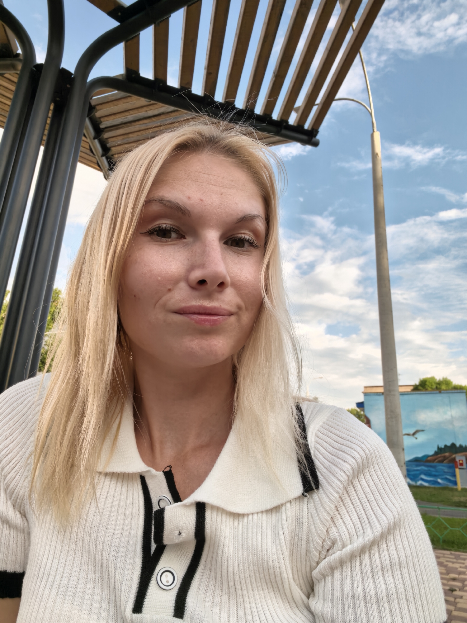 A young woman with blonde hair and light skin is taking a selfie outdoors. She is wearing a white, ribbed polo shirt with black trim and buttons. She is seated under a canopy with wooden slats and a metal frame. In the background, there is a blue sky with scattered clouds, a lamppost, and a building with a mural depicting a seascape with a bird. The image appears to be taken in Sterlitamak, Russia.